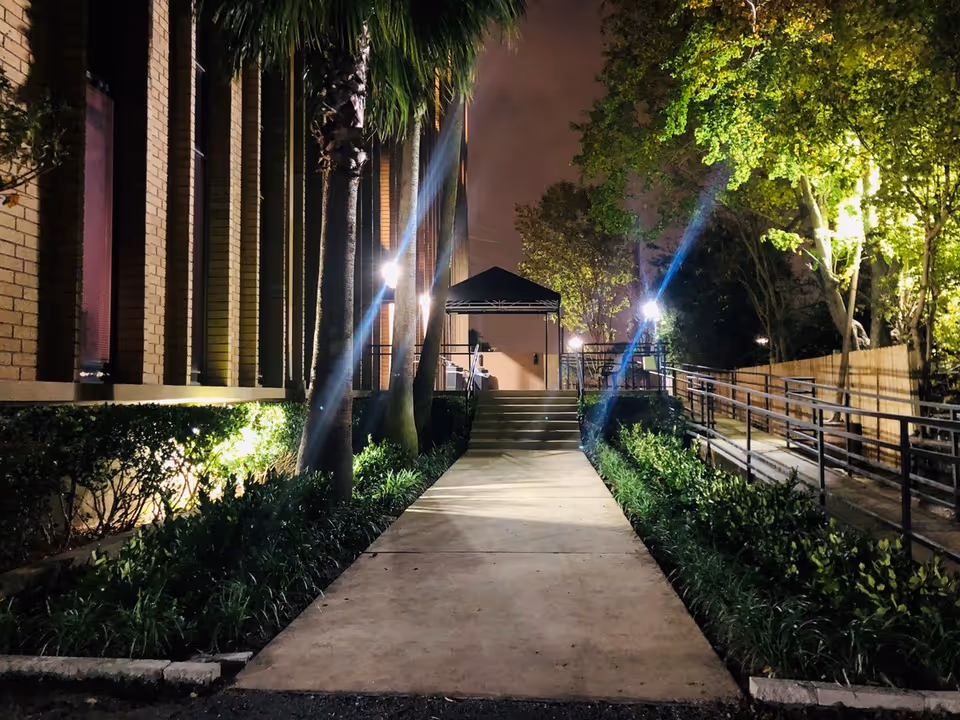 Night exterior view of a lit walkway with stairs and landscaped sides to the entrance.