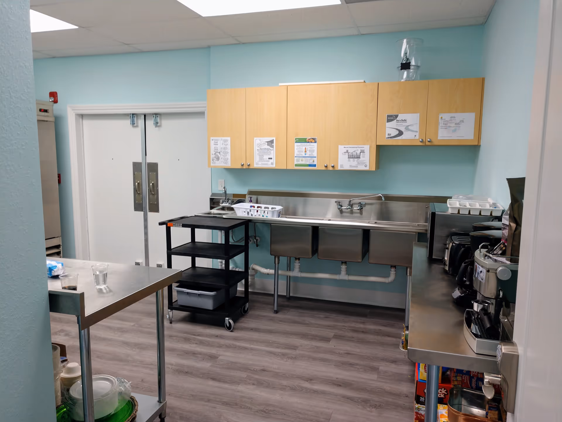 Commercial kitchen with stainless sinks, cabinets, and appliances in a light blue-walled room.
