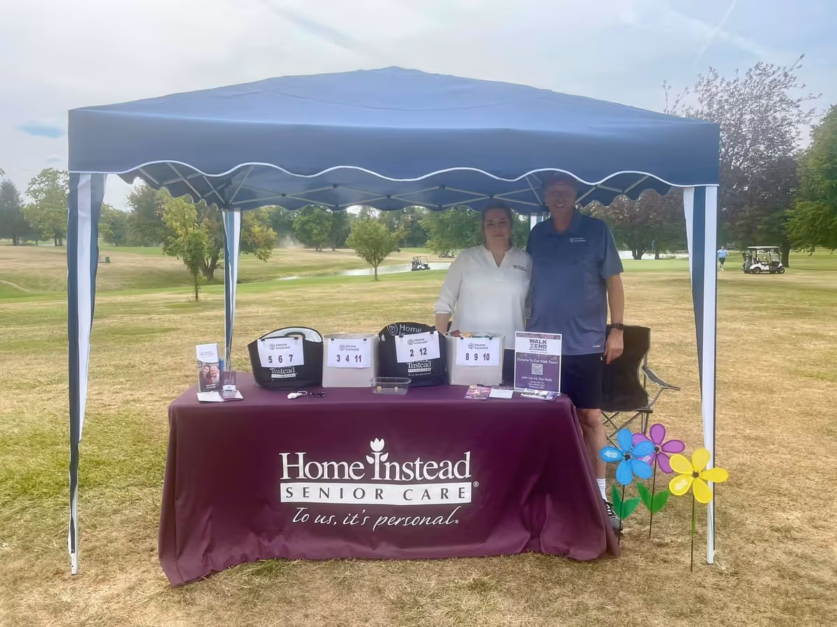 Two staff members stand behind a maroon Home Instead table under a blue canopy outdoors.