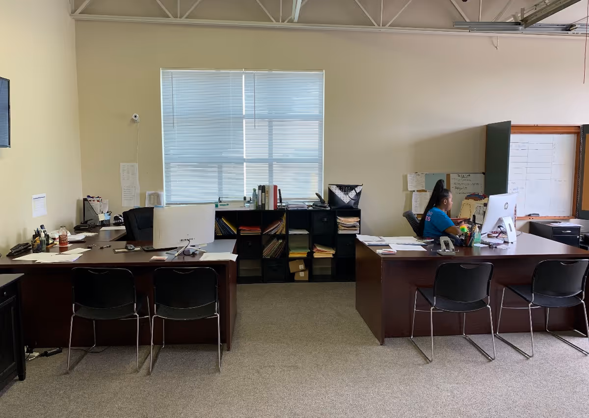 Office reception area with two desks, chairs, files, and a person working at a computer.