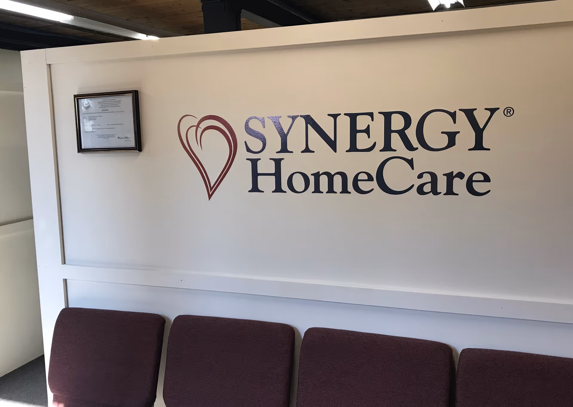 An interior lobby area with a SYNERGY HomeCare sign and a row of maroon seats.
