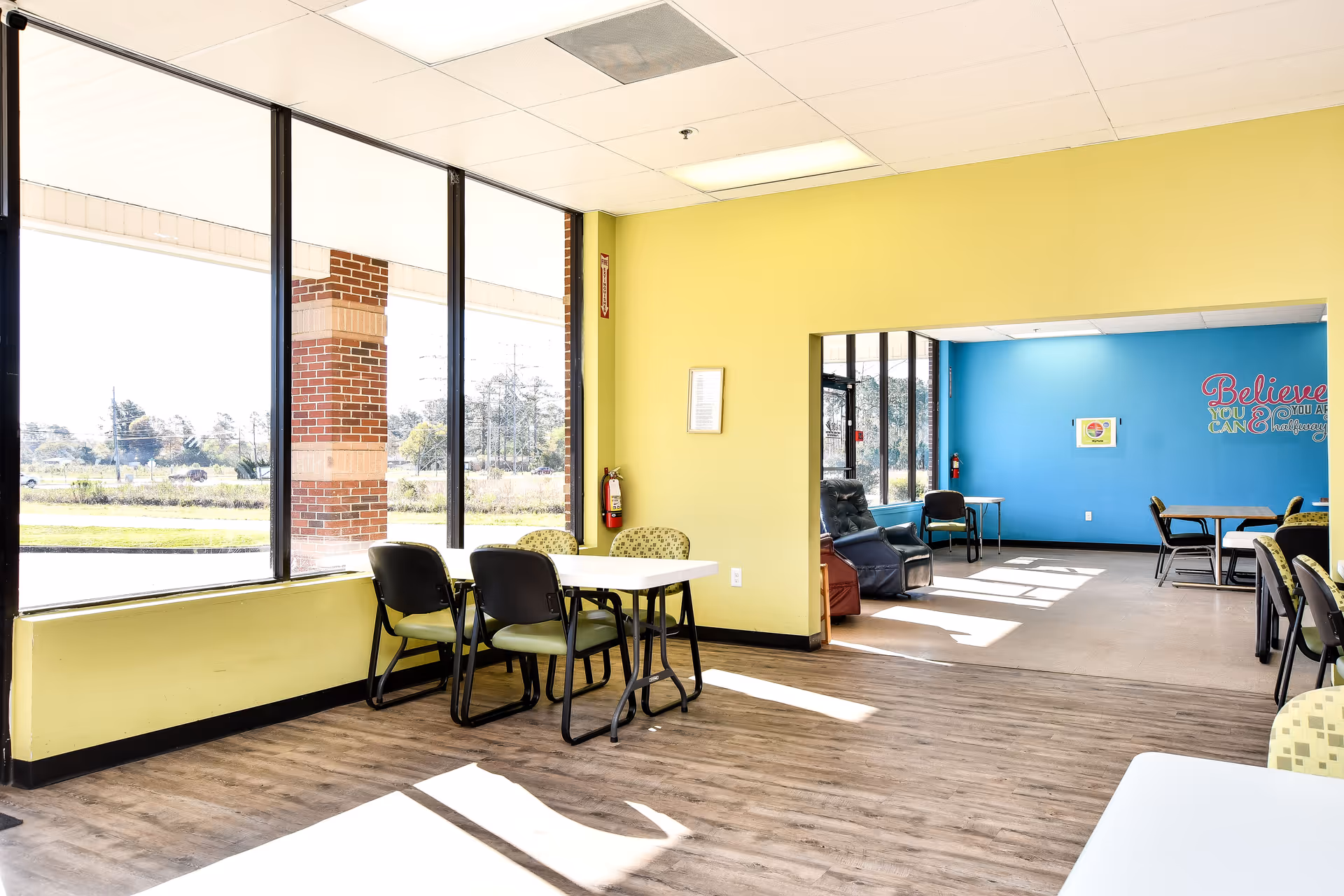 Bright communal lounge area with tables, chairs, and large windows.