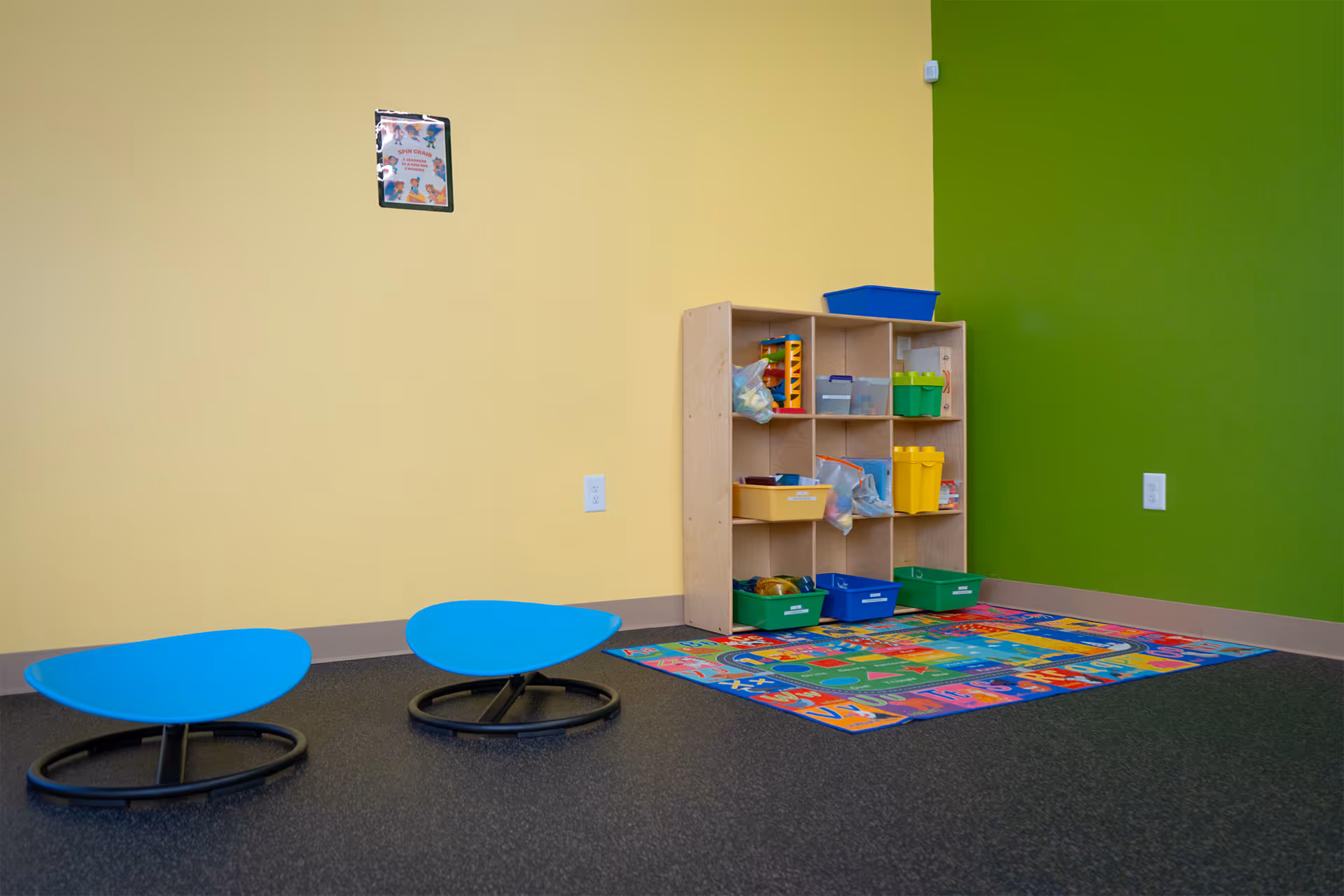 Bright pediatric therapy room with yellow and green walls, a cubby shelf, rug, and blue seats.