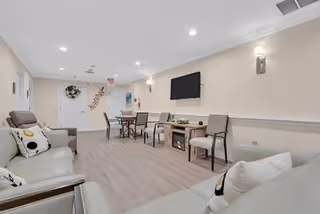 Bright senior living lounge with sofas, chairs, tables, and a wall-mounted TV.