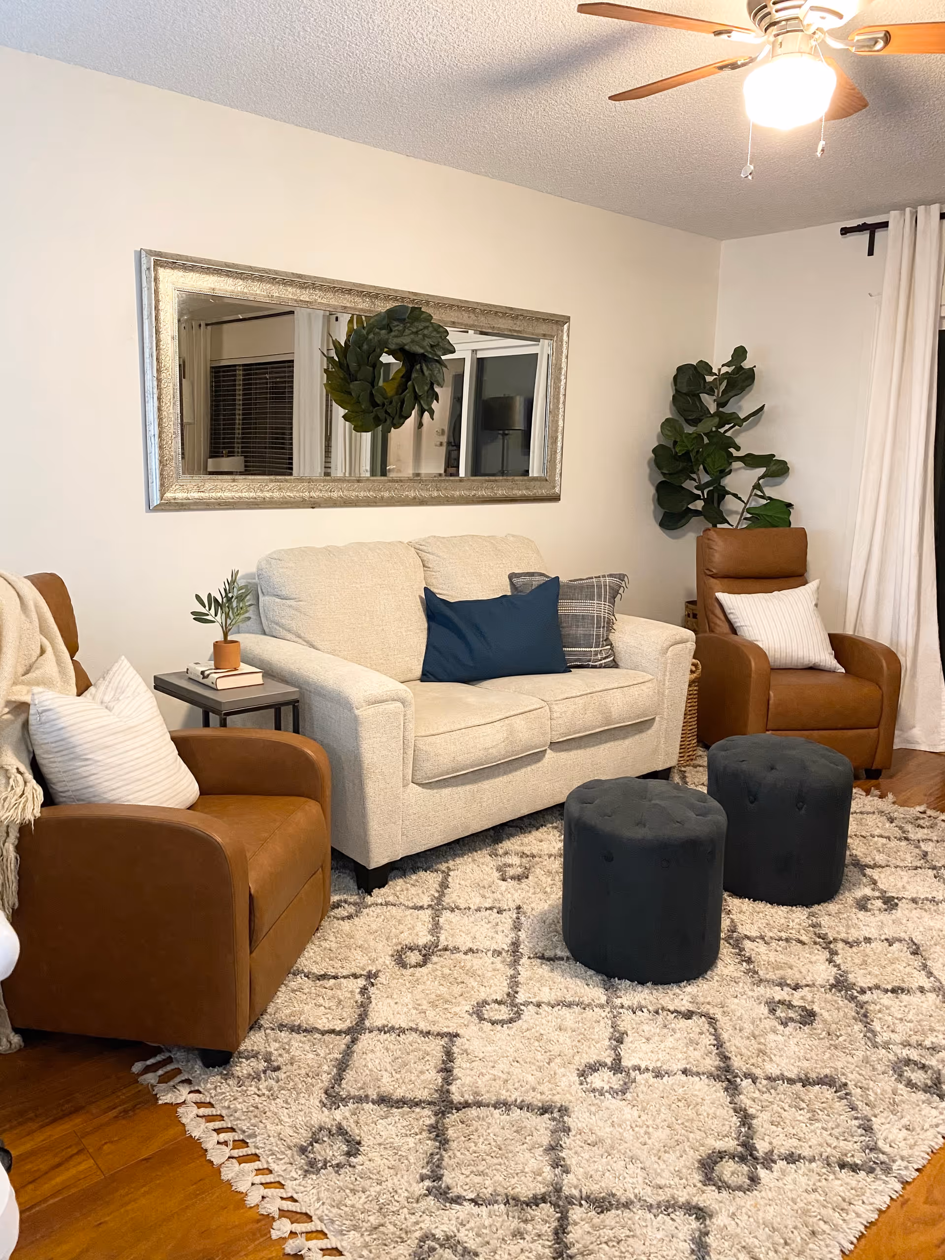 Cozy living room with beige sofa, brown chairs, patterned rug, plants, and a wall mirror.