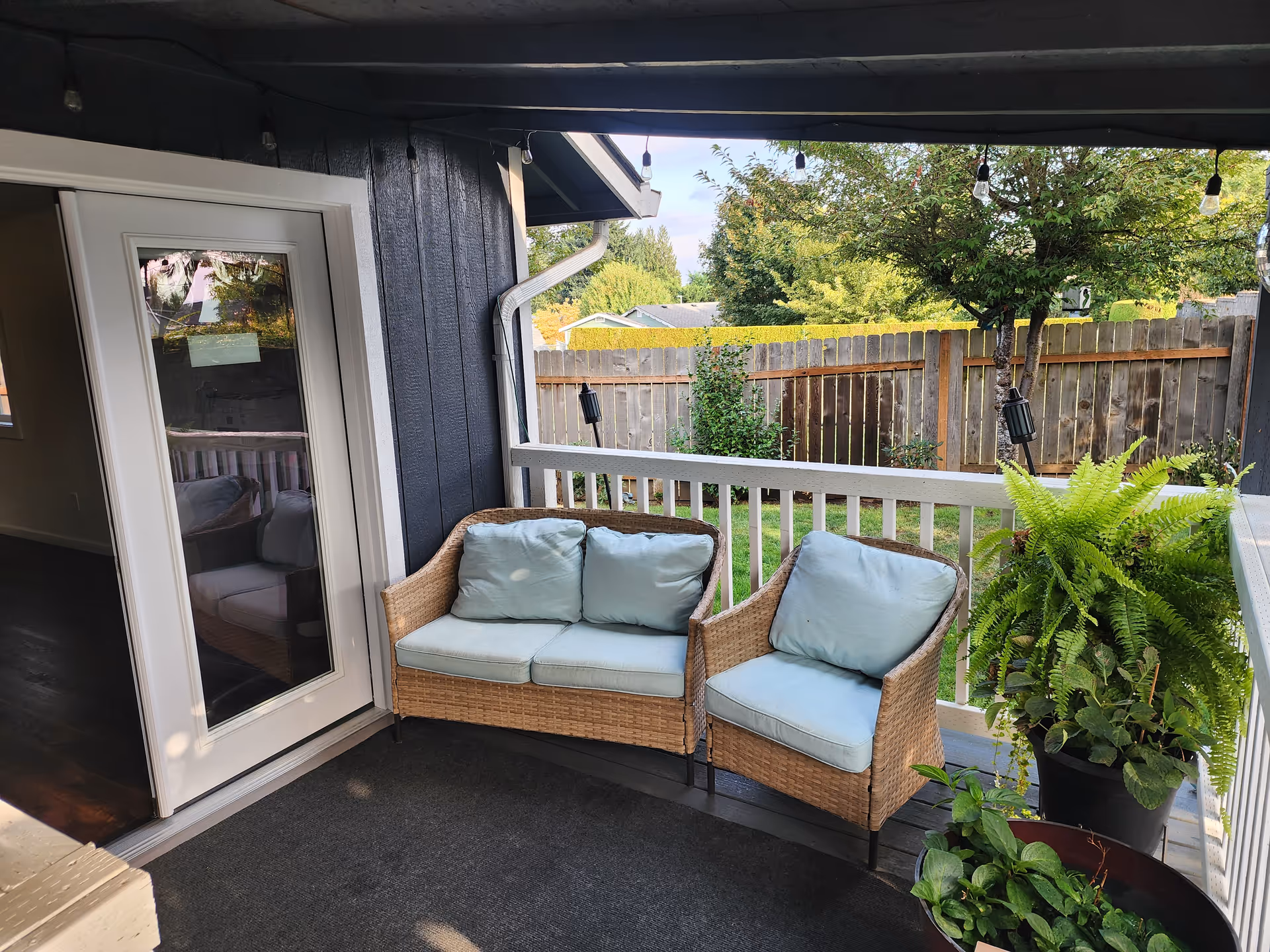 Outdoor balcony with wicker seating, blue cushions, potted plants, string lights, and a wooden fence.