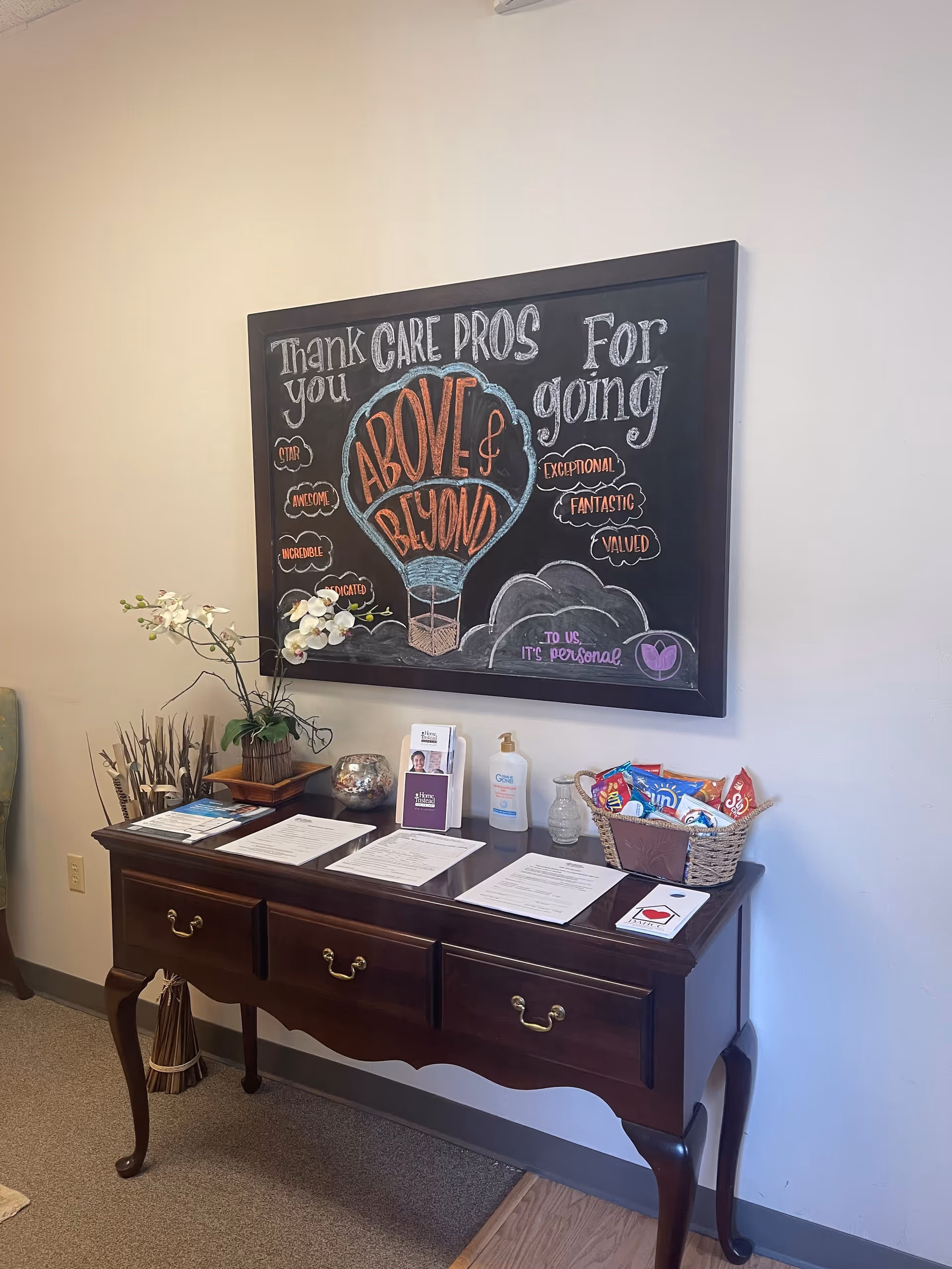Chalkboard art above a wooden console with papers, sanitizer, flowers, and a snack basket in hallway.