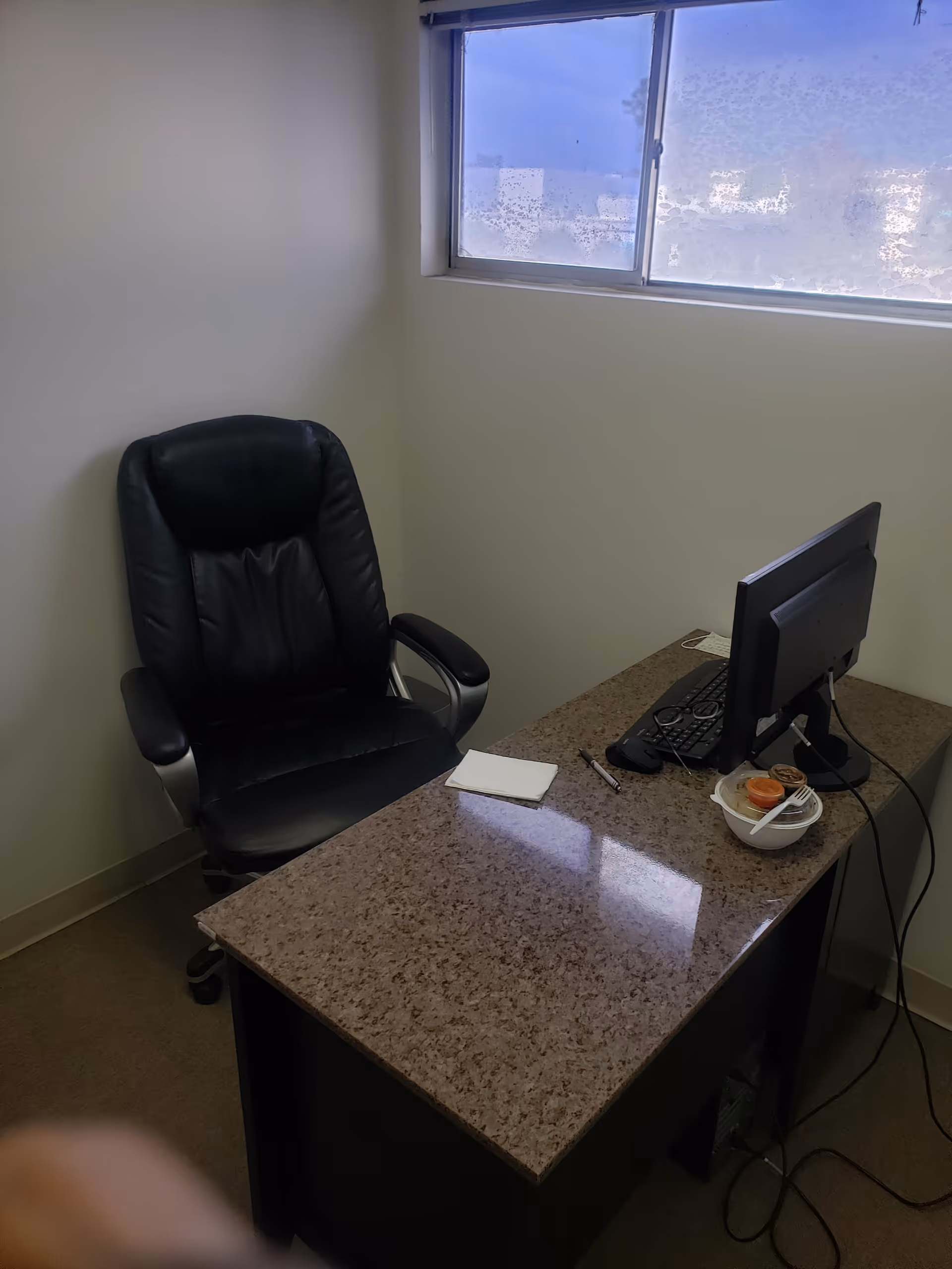 Compact office featuring a granite desk, black leather chair, and computer by a window.