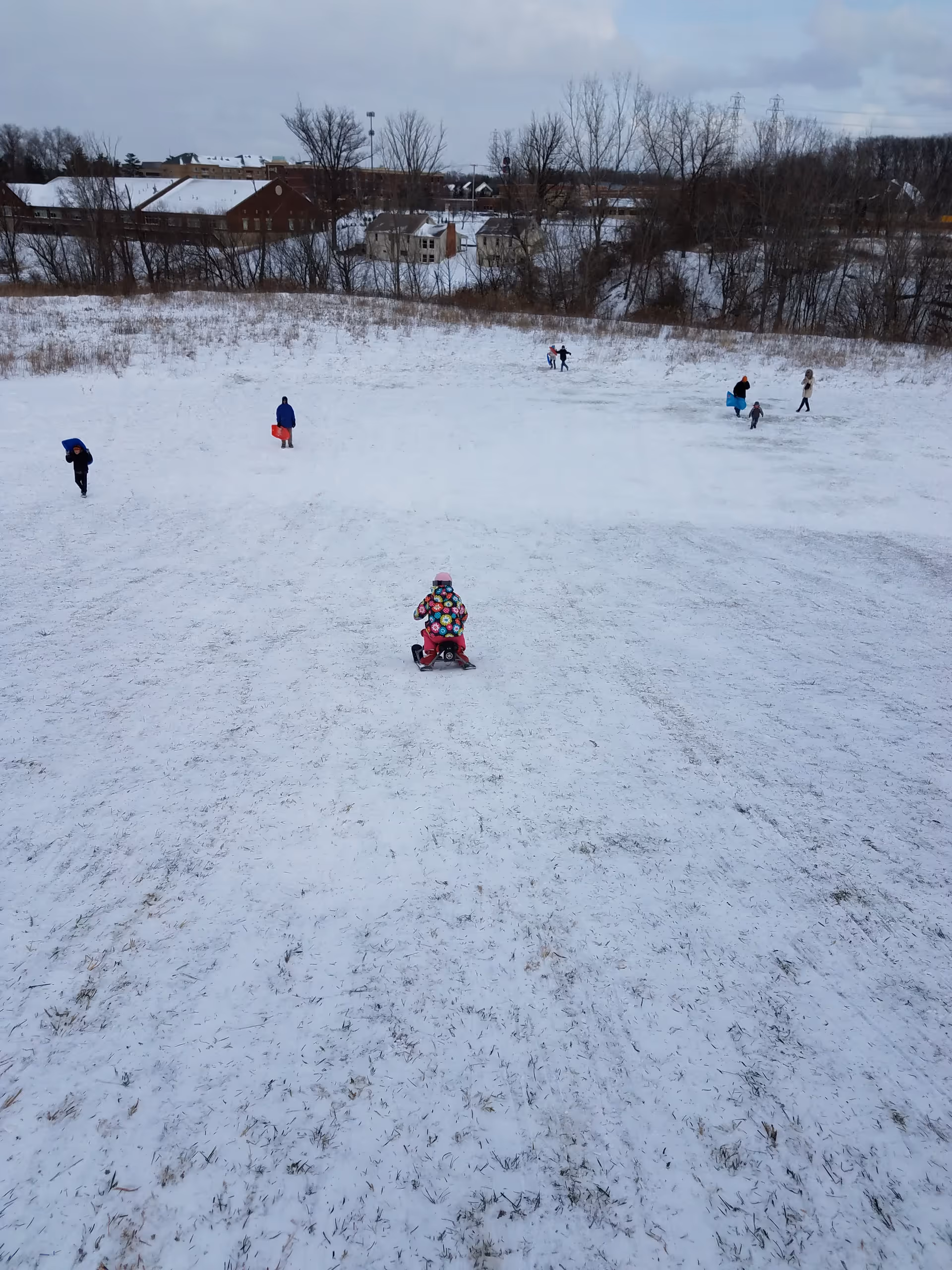 Snowy open field with children playing; foreground child on a pink snow scooter.