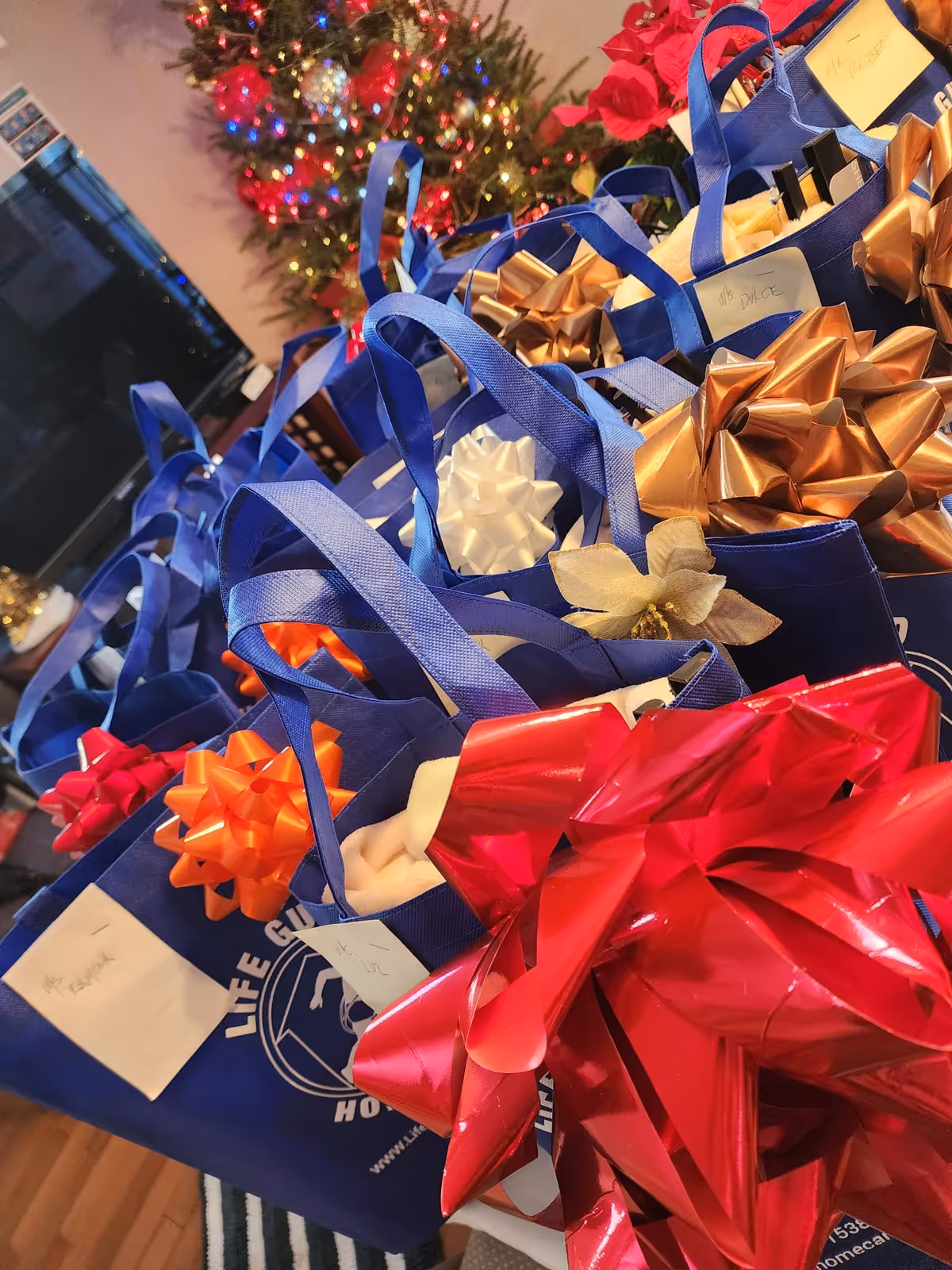 Blue gift bags with ribbons and tags under a Christmas tree indoors.