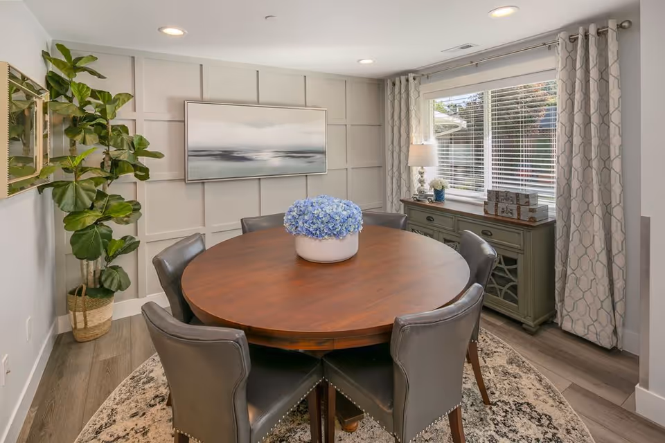 Round wooden dining table with six leather chairs, blue floral centerpiece, and windowed sideboard.