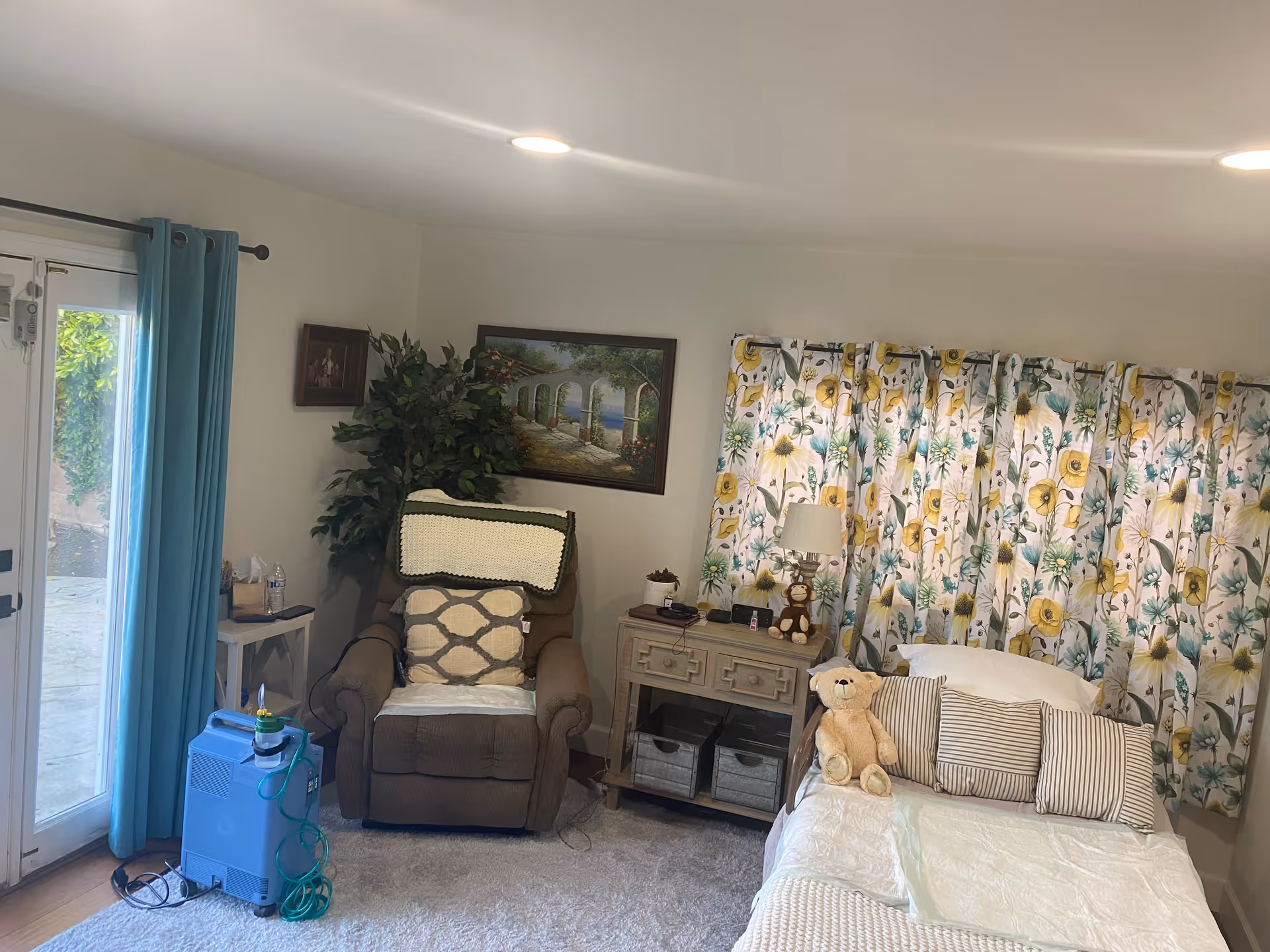Cozy bedroom with a bed, recliner chair, floral curtains, lamp, and a teddy bear.