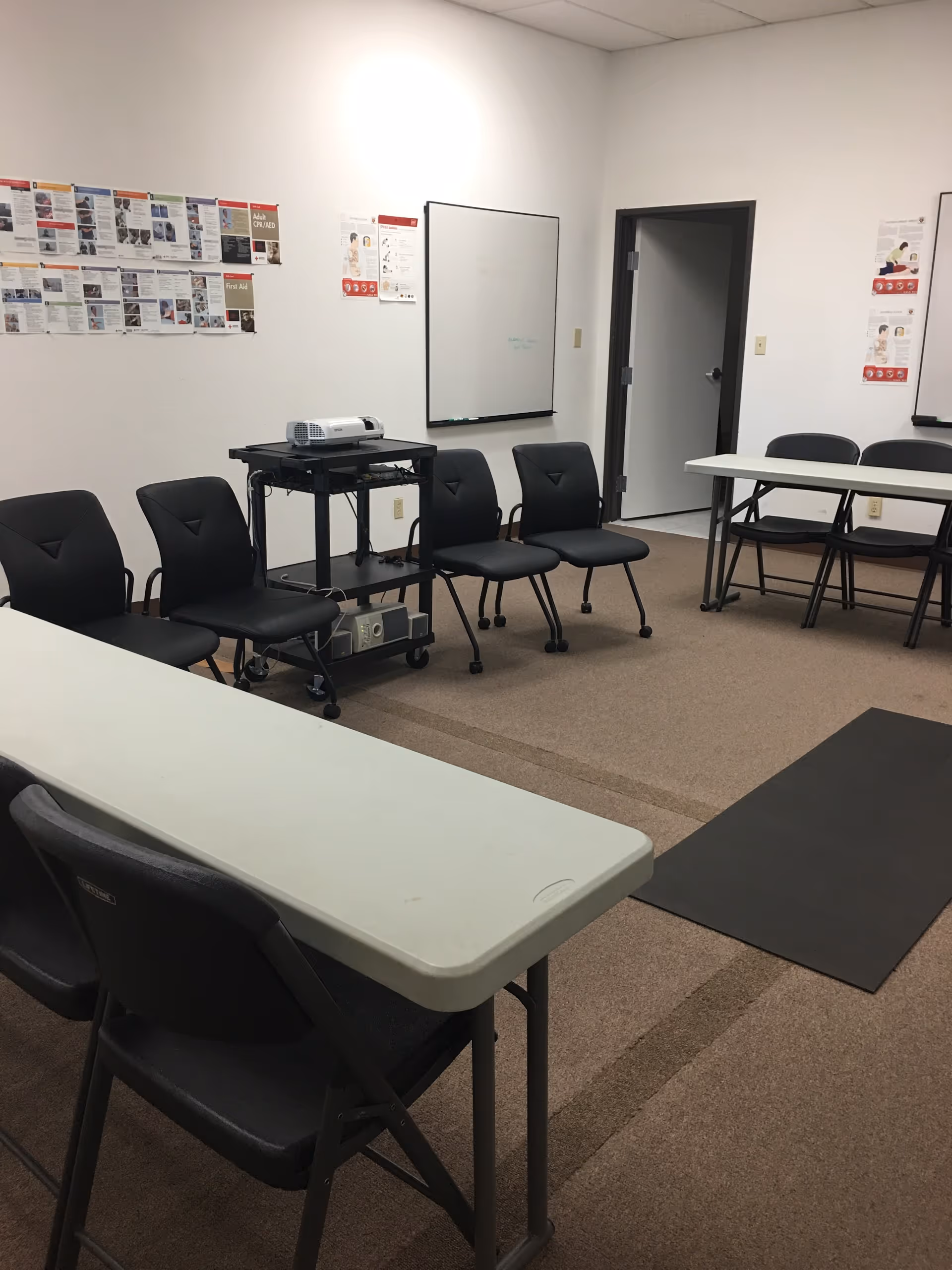 A training room with folding tables, black chairs, a projector cart, whiteboard, and posters.
