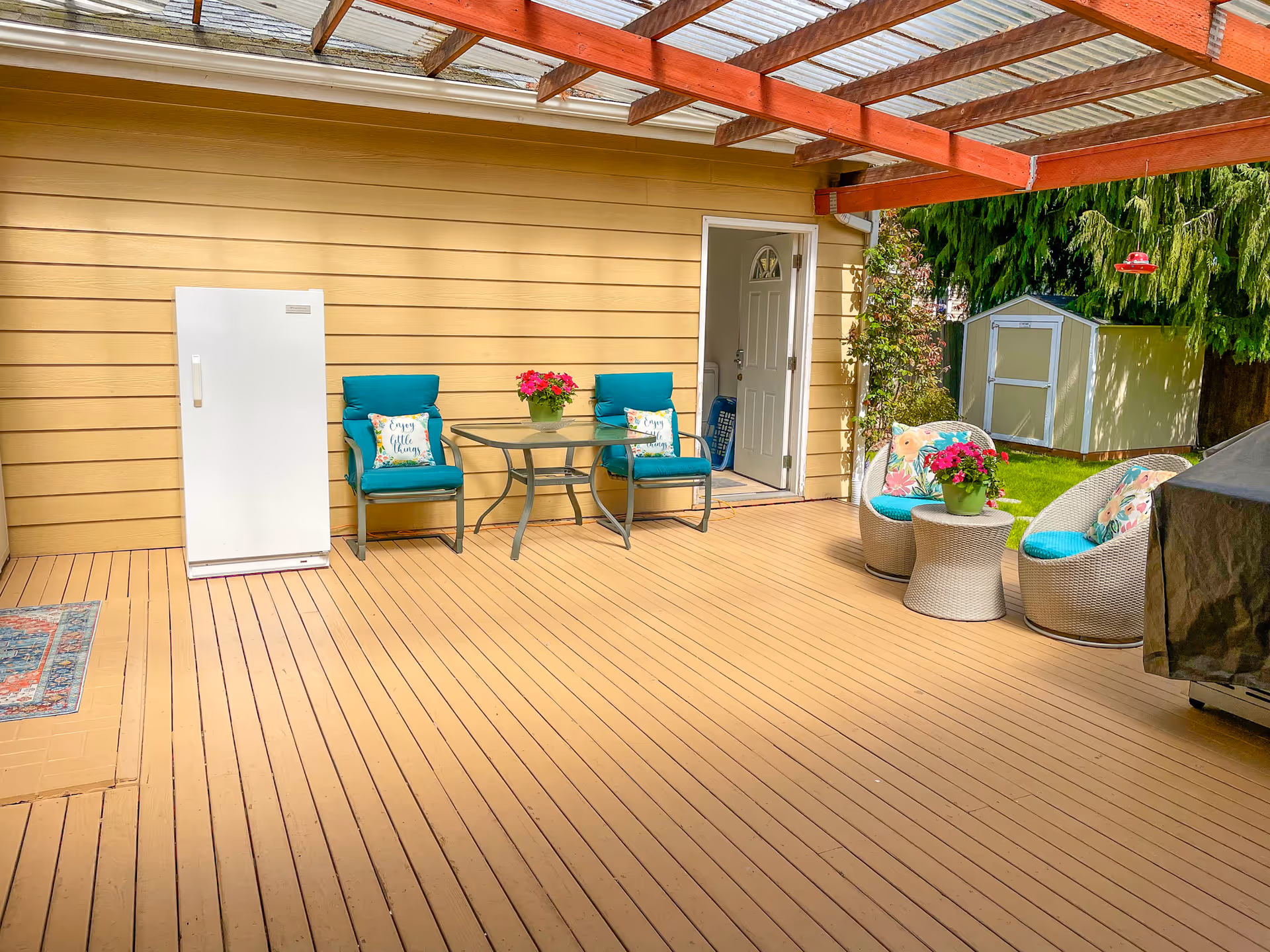 Sunny outdoor deck with turquoise cushioned seating, a glass table, potted plants, and an entry door.