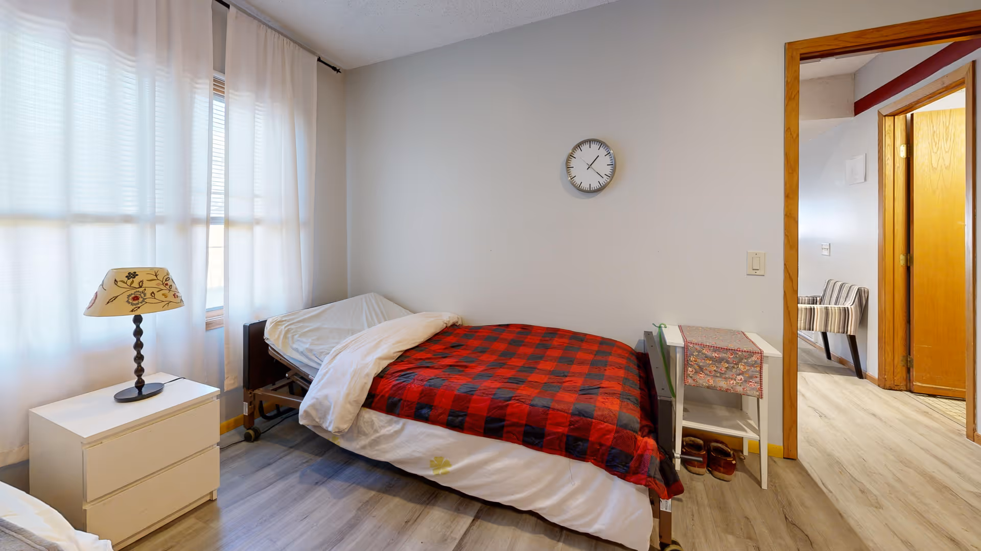 A small senior living bedroom with a single bed, plaid blanket, lamp, window, and clock.