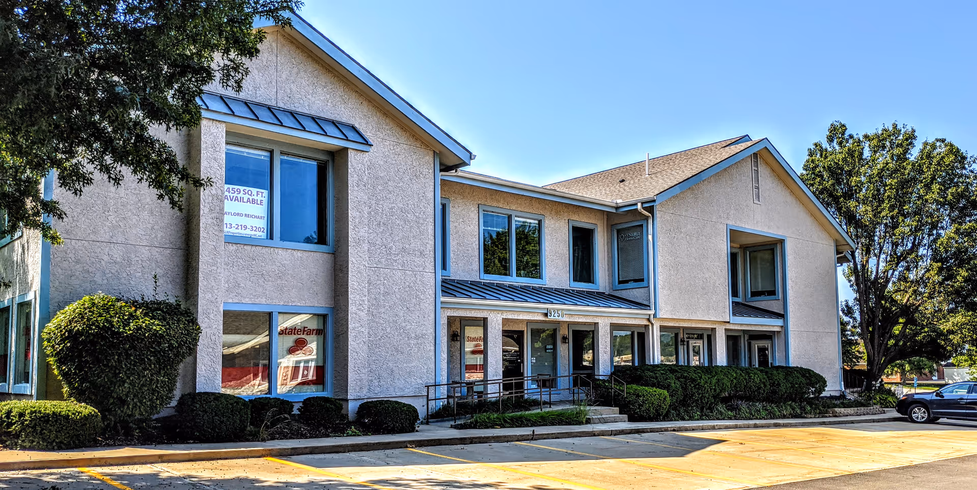 Two-story beige building with large windows, manicured shrubs, and a parking lot under a clear sky.