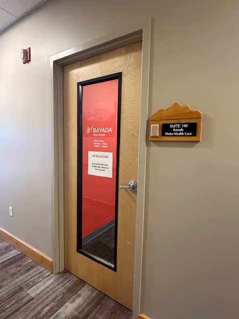 Interior hallway with a wooden door featuring glass panel and BAYADA Home Health suite plaque.