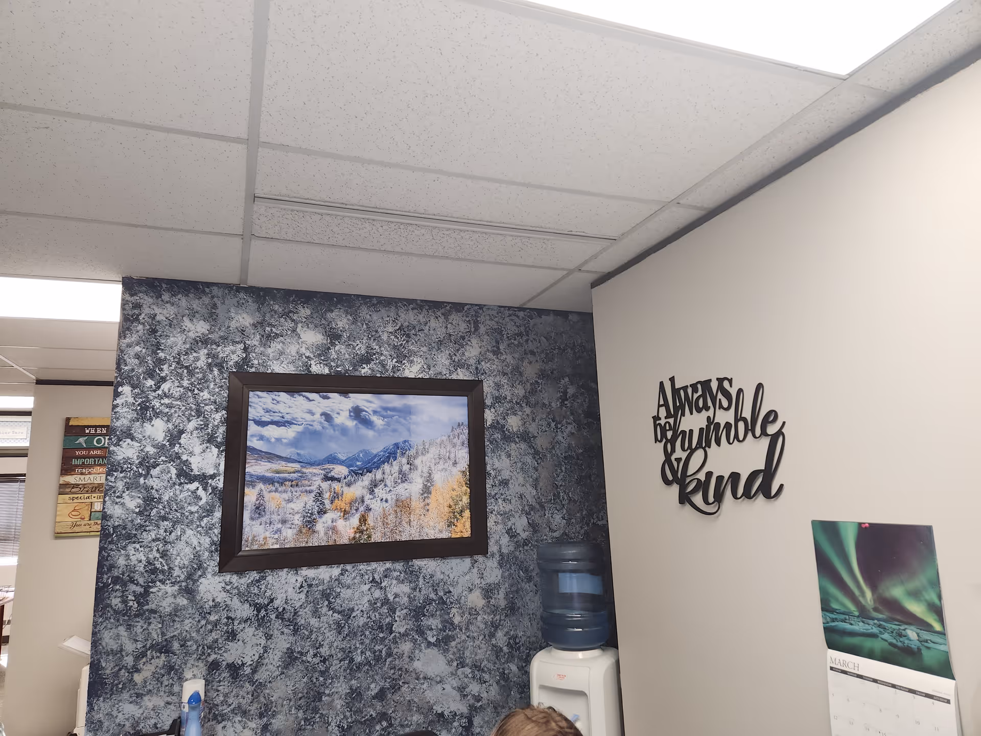 Indoor common area with textured blue wall, framed landscape, water cooler, and inspirational wall art.