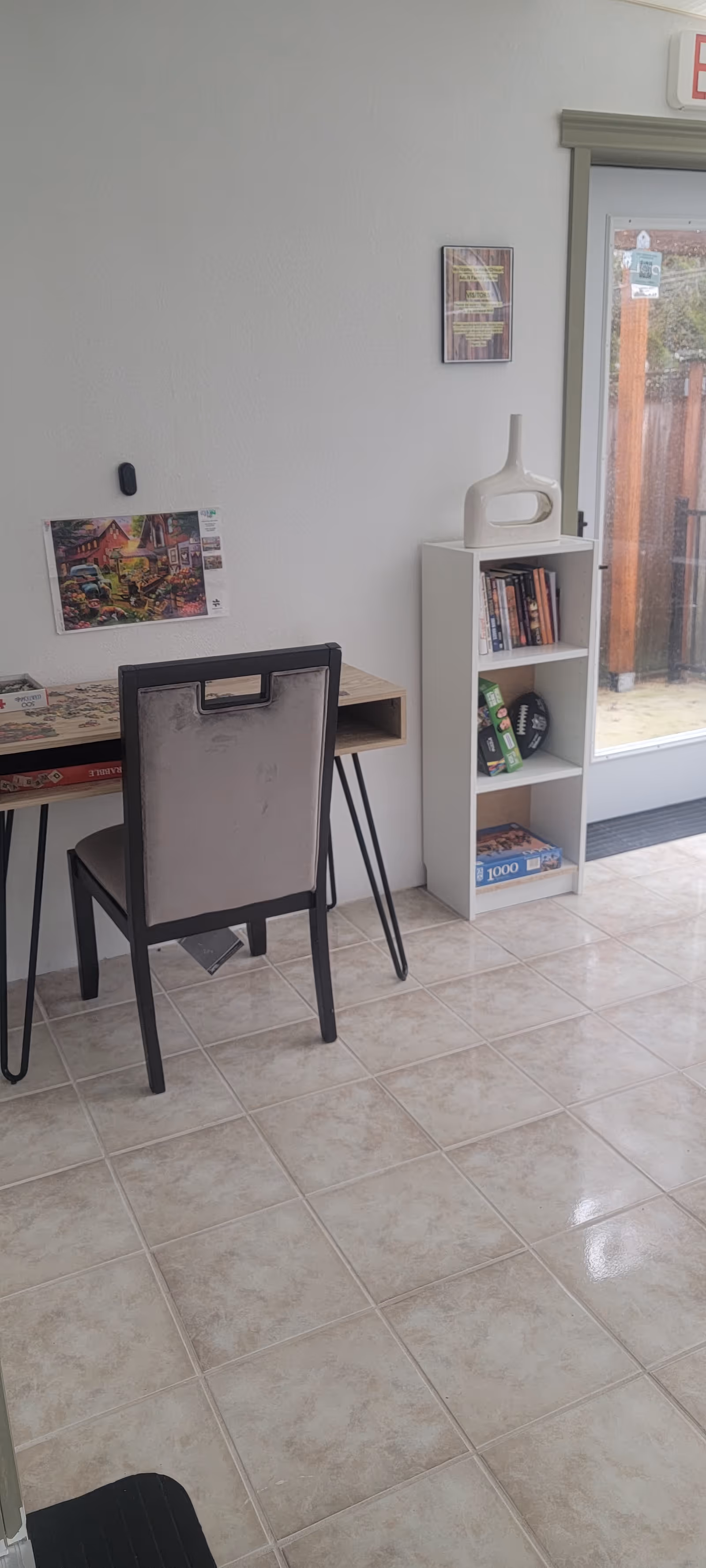 Interior view of a small room with a desk, chair, bookshelf, and exit door.