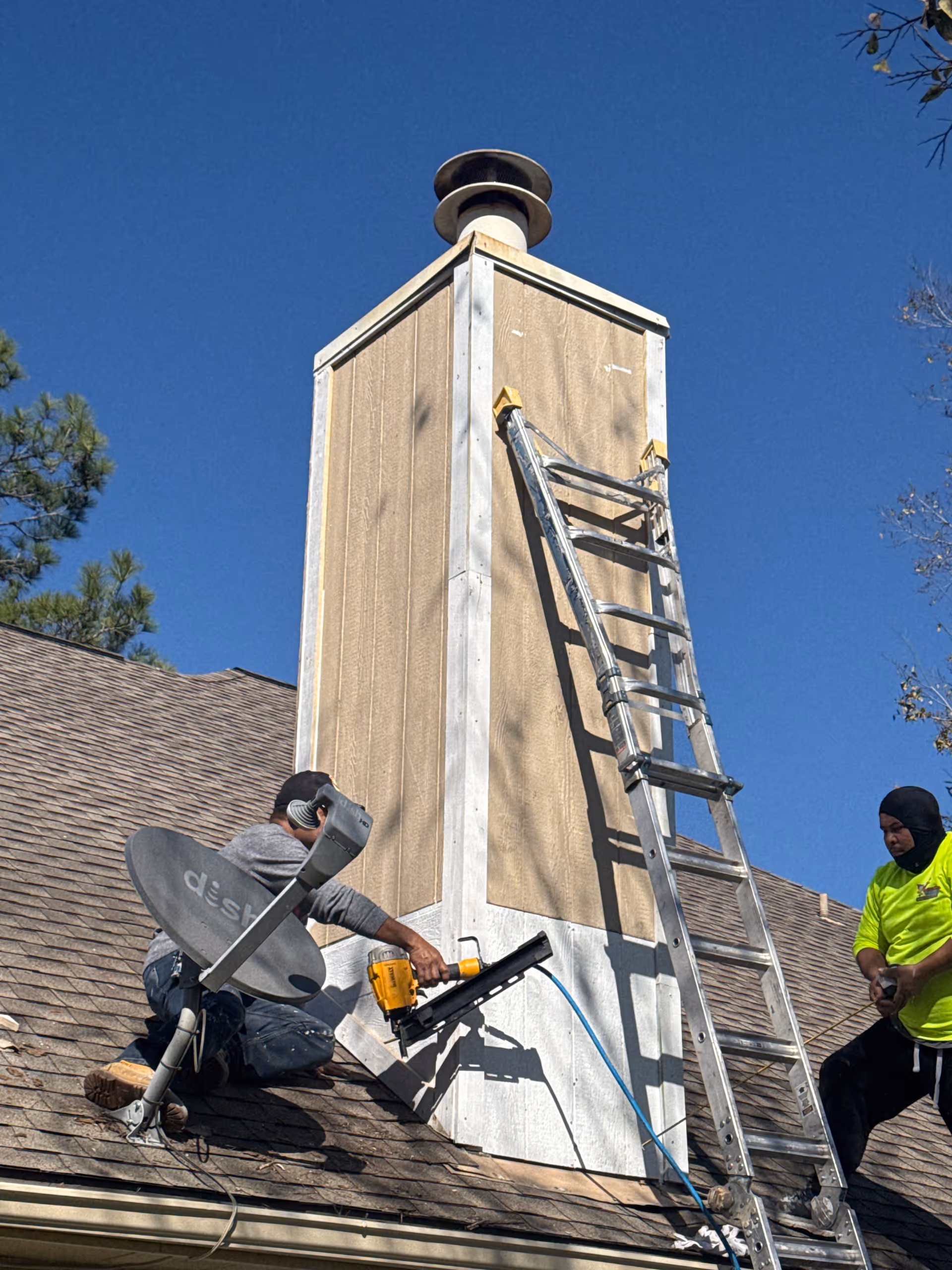 Two workers on a sloped roof repairing a chimney with a ladder nearby.