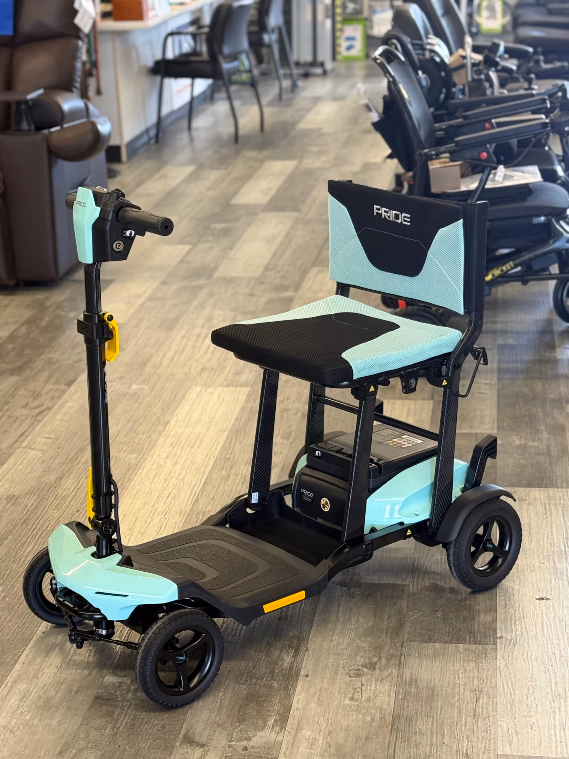 Mint-green mobility scooter on a showroom floor with chairs visible in background.