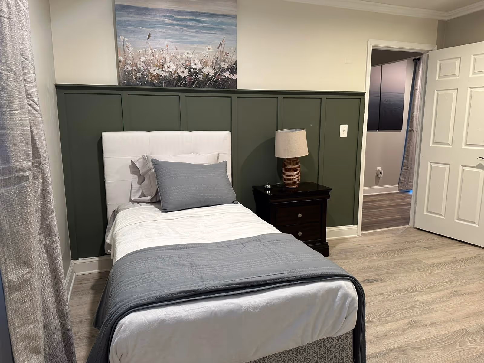 A tidy bedroom with a white bed, gray bedding, green paneled wall, and a nightstand.