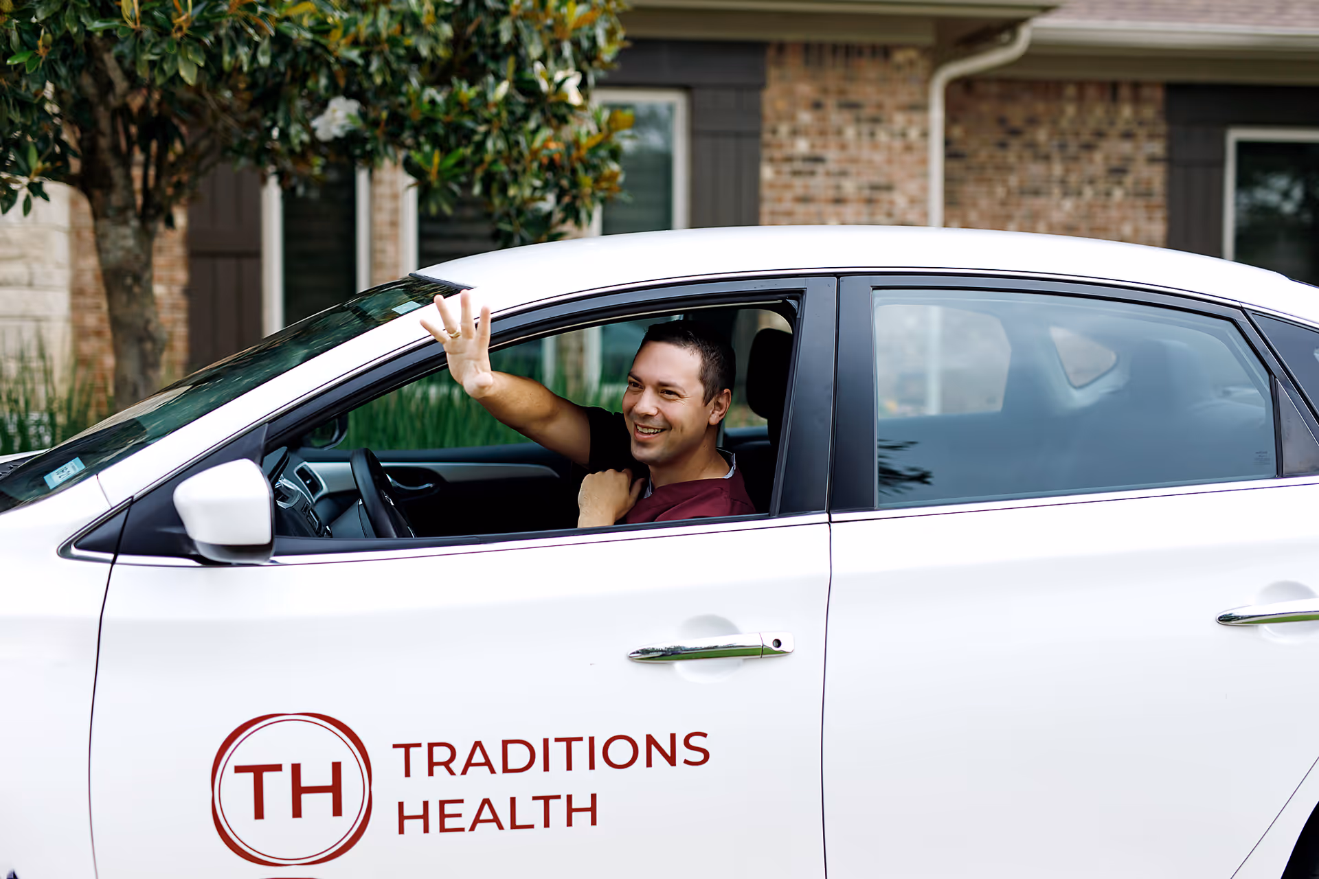 Person waves from a white Traditions Health vehicle parked in front of a house.