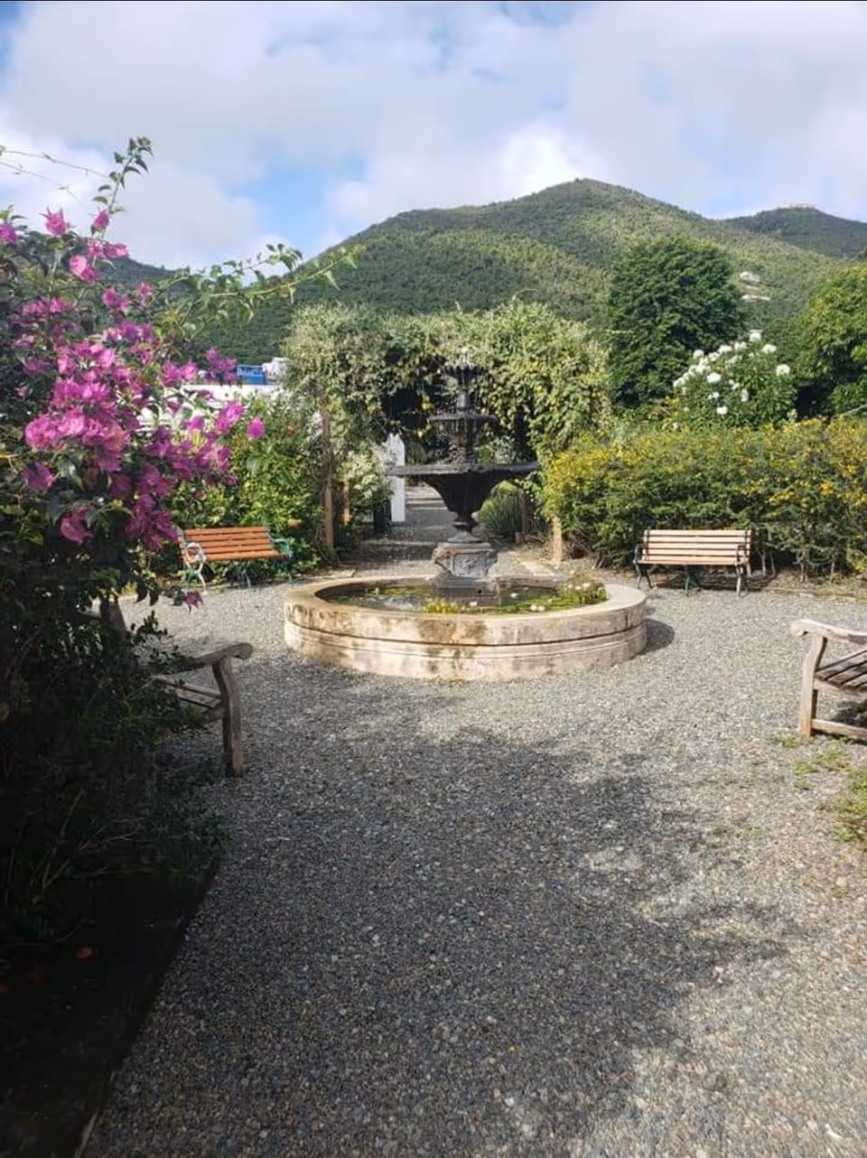 An outdoor courtyard with a central fountain, gravel path, benches, and flowering shrubs.