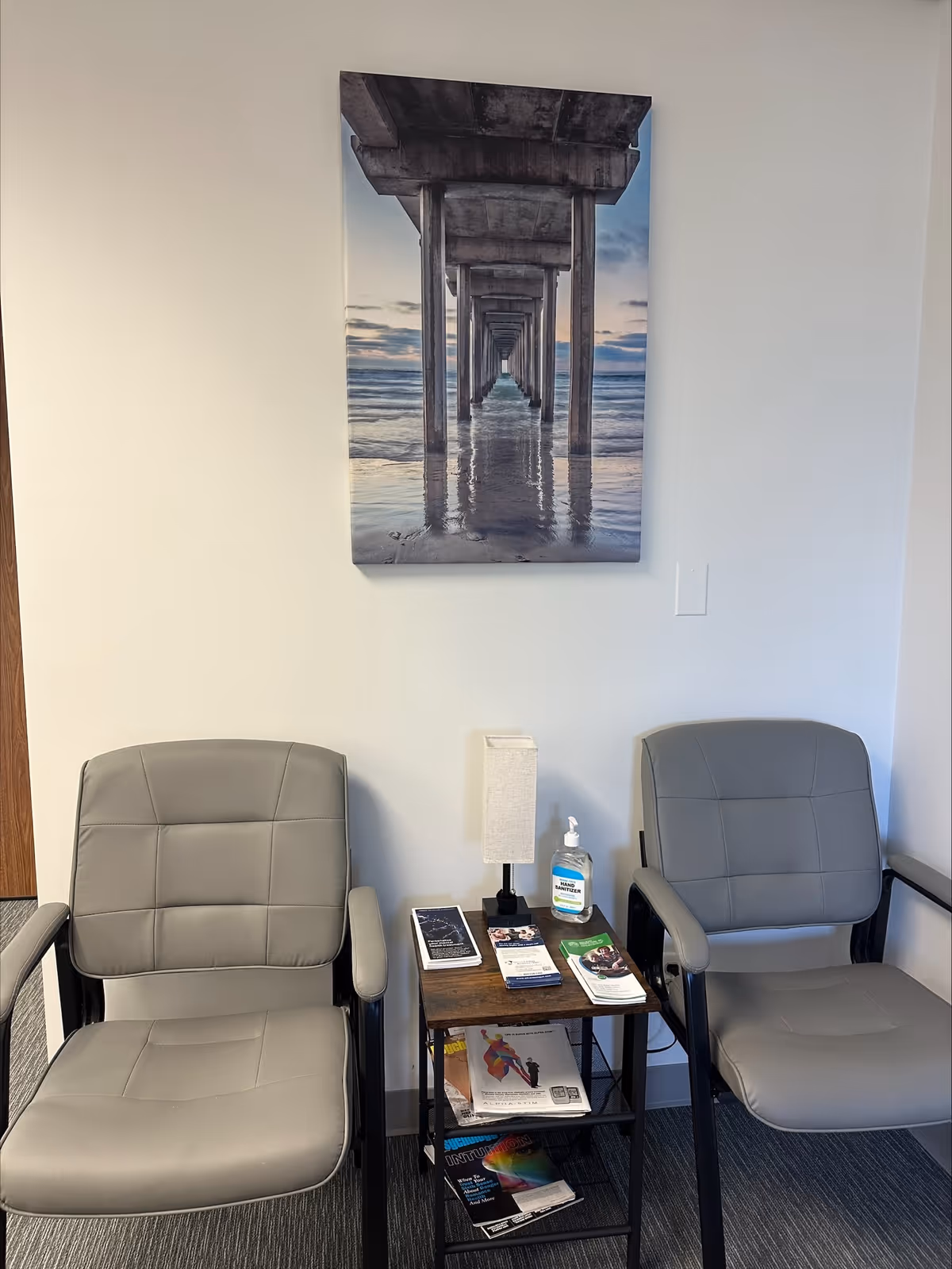 Two gray chairs sit beside a small table with brochures, hand sanitizer, and a pier photograph.