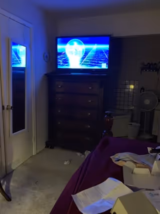 A bedroom with a dresser, a TV on top, a purple bedspread, and scattered papers.