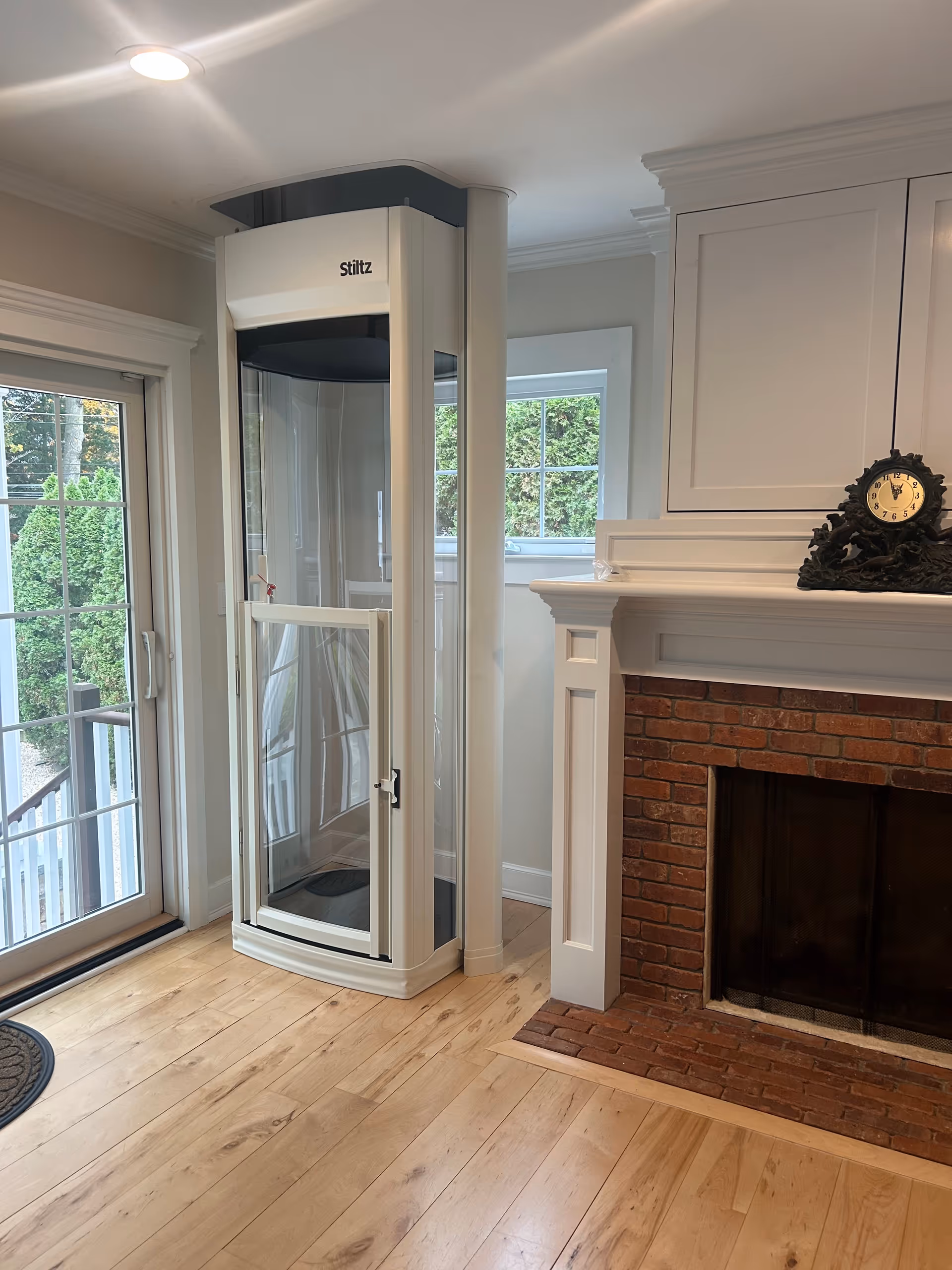 A white residential elevator stands beside a brick fireplace in a bright living room.