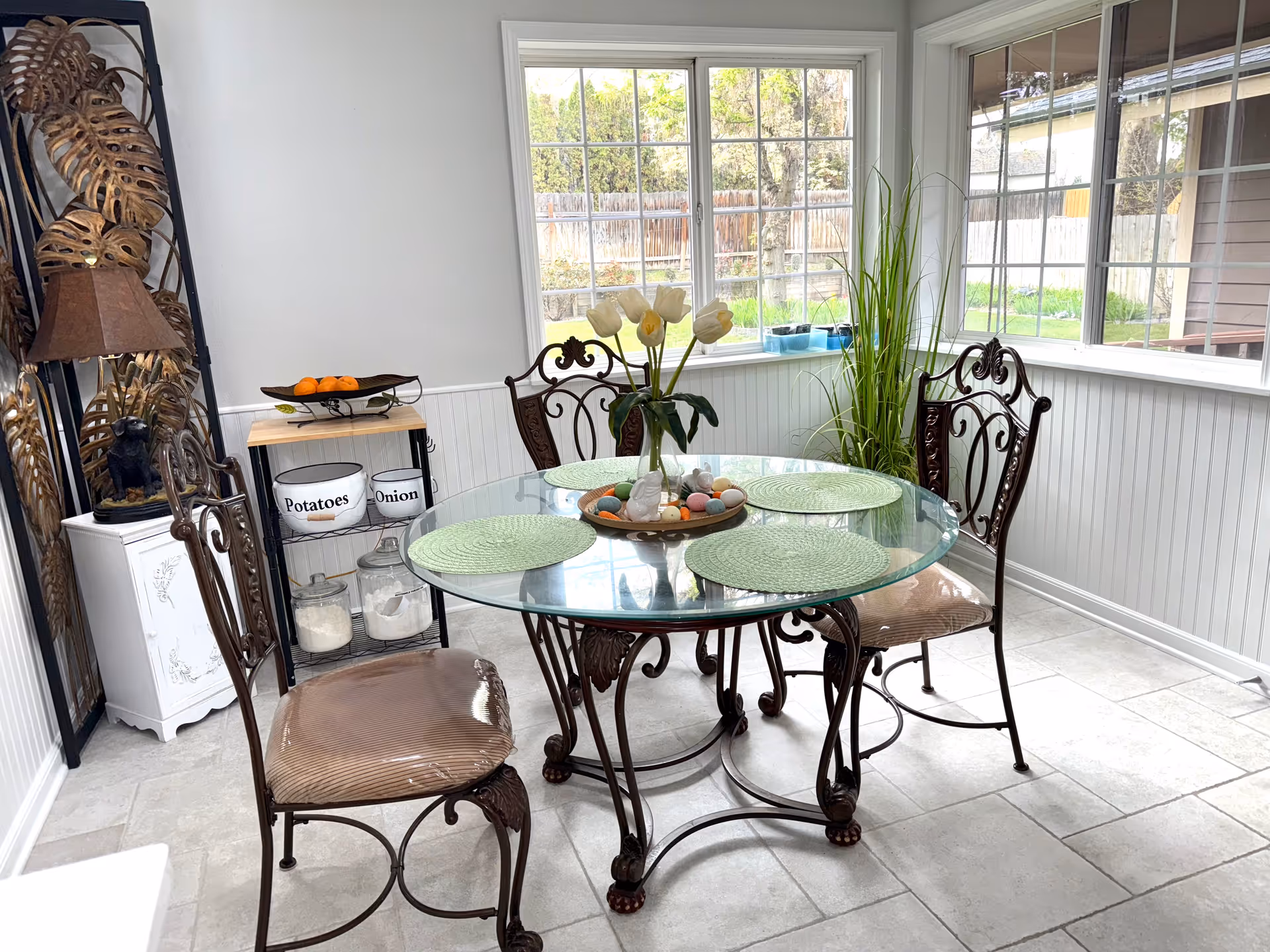 Sunlit dining room featuring a glass round table, ornate chairs, and tall plants near windows.