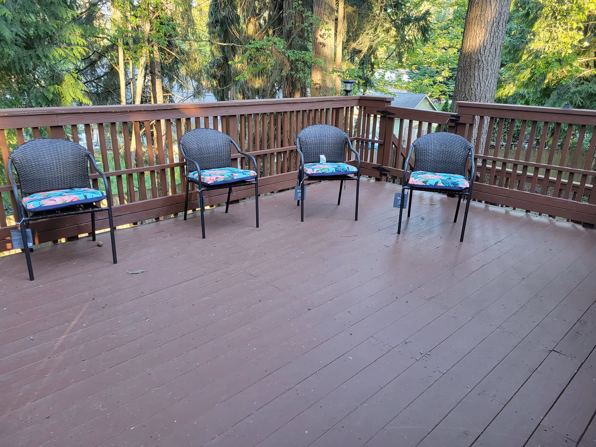 Outdoor wooden deck with five chairs and colorful cushions, surrounded by trees.