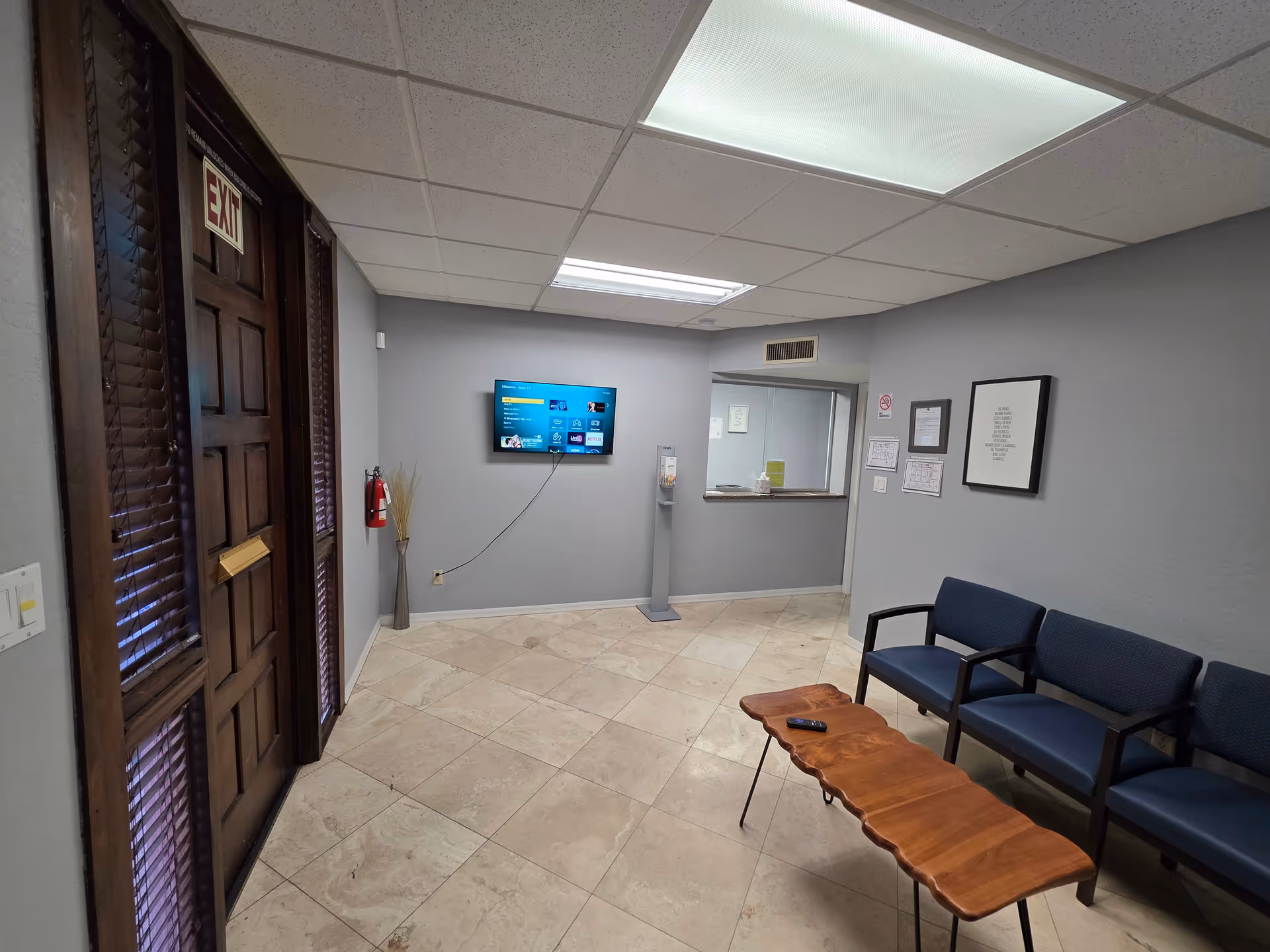 Interior waiting area with blue chairs, a wooden table, and a wall-mounted TV.