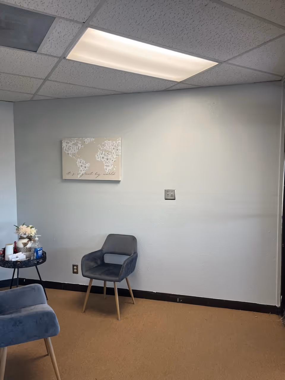 A small waiting area with blue chairs, a side table, flowers, and world map wall art.