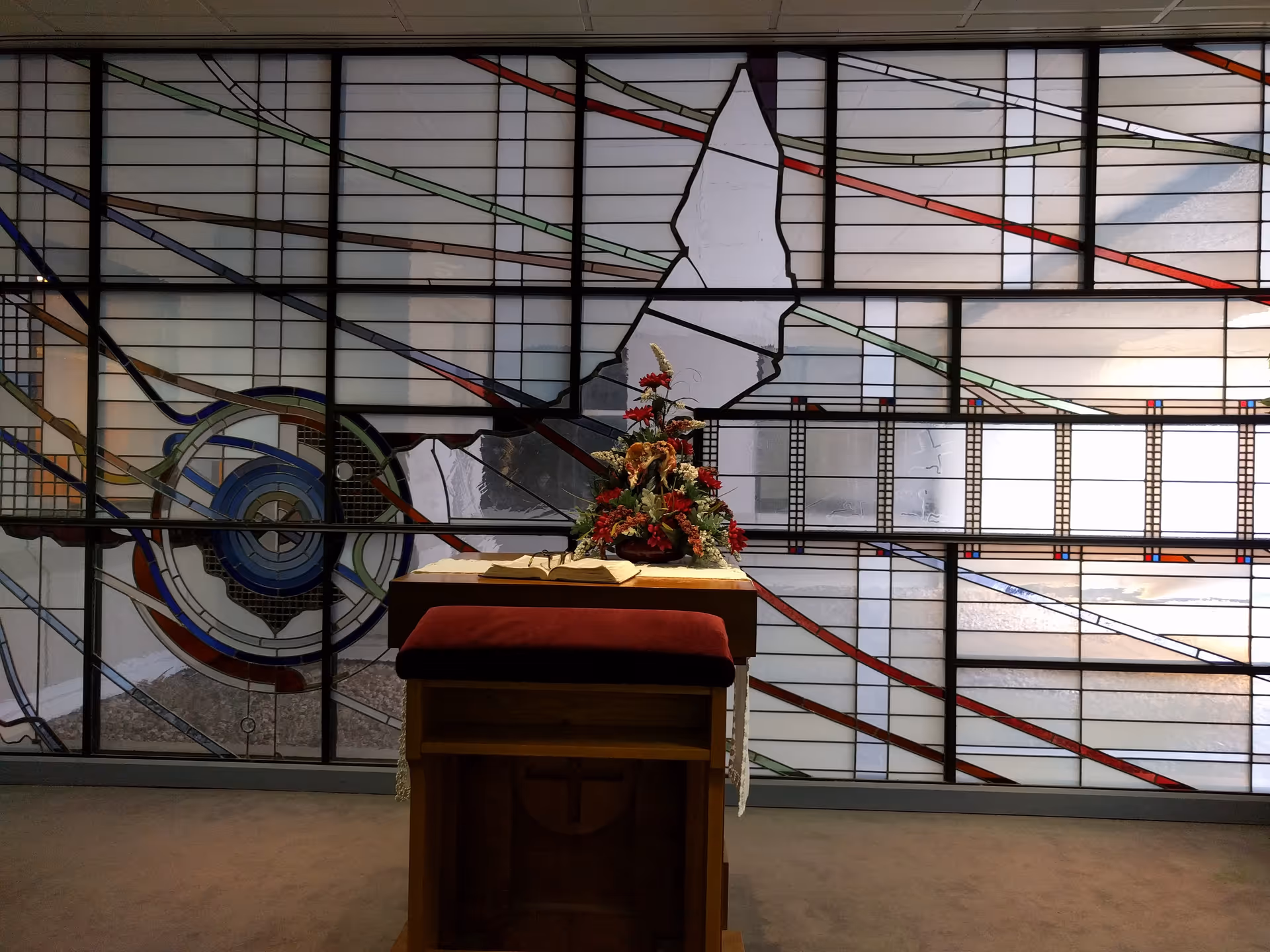 A wooden lectern with flowers stands before a large stained-glass wall indoors.
