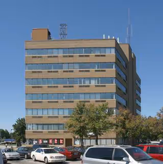 Tall multi-story beige building with numerous windows, surrounding parking lot and a clear blue sky.