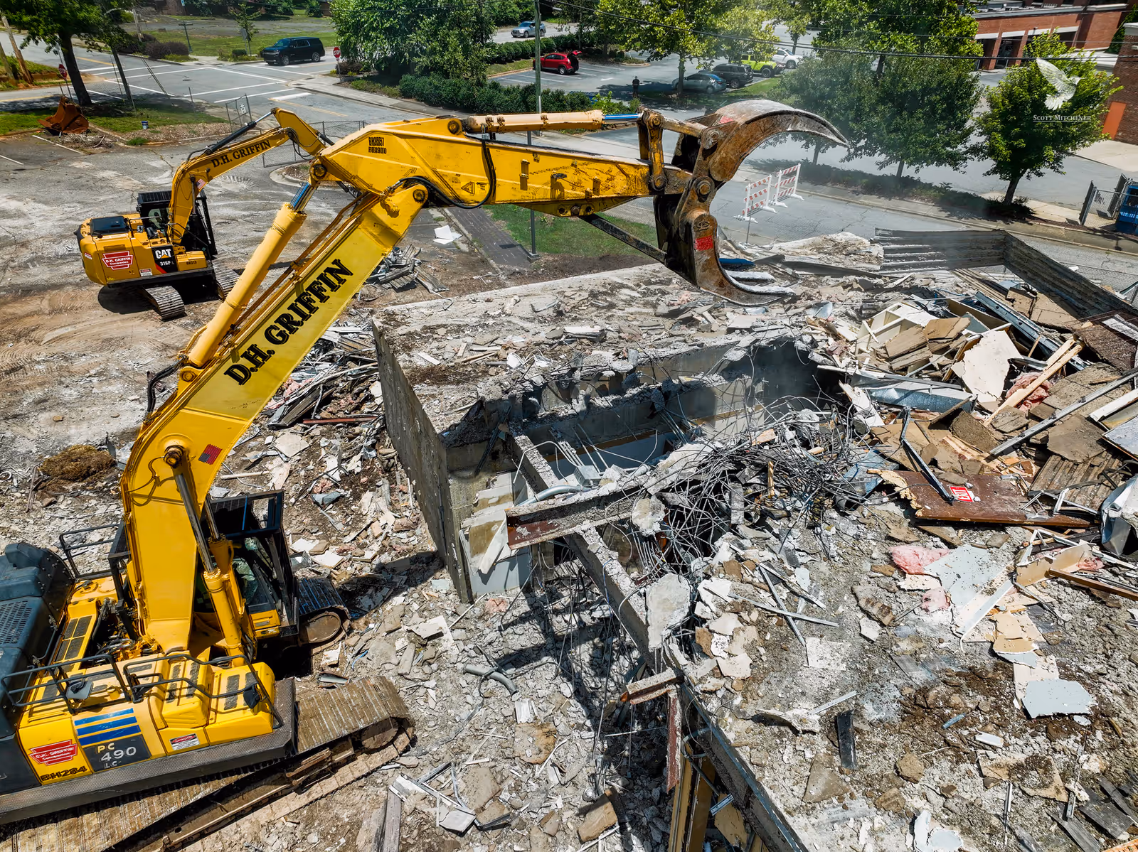 A large yellow excavator demolishes a building on a rubble-filled site.