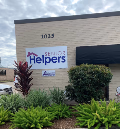 Exterior of a Senior Helpers facility with sign, beige brick wall, and landscaped plants.