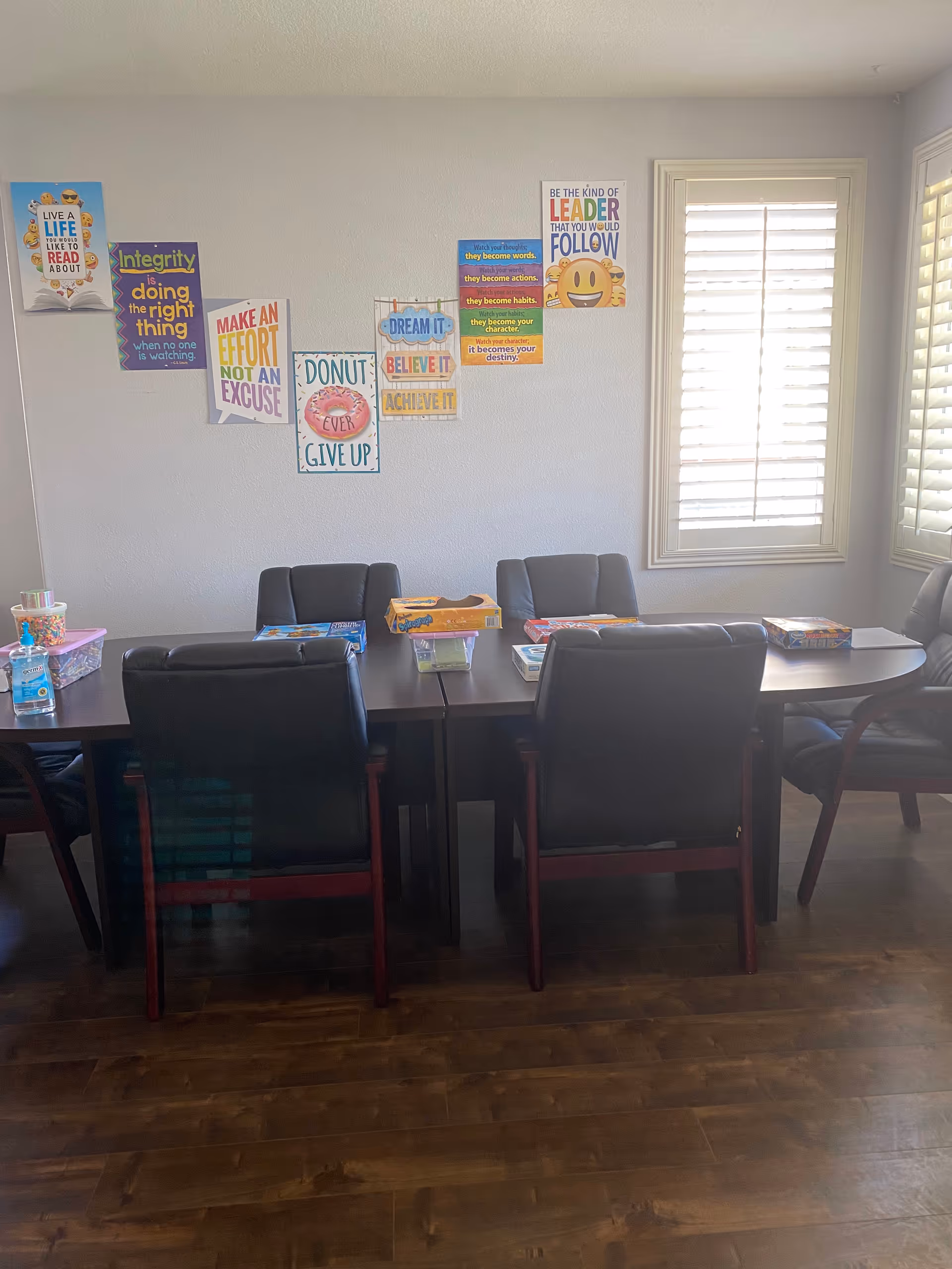Communal dining room with a long table, chairs, posters on the wall, windows, and wooden floor.