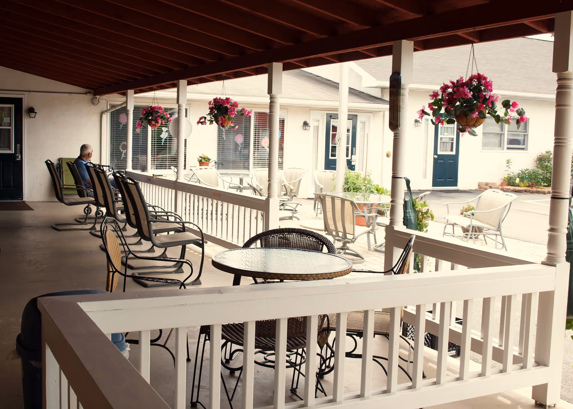Covered porch with chairs, tables, hanging baskets, and a resident seated nearby.