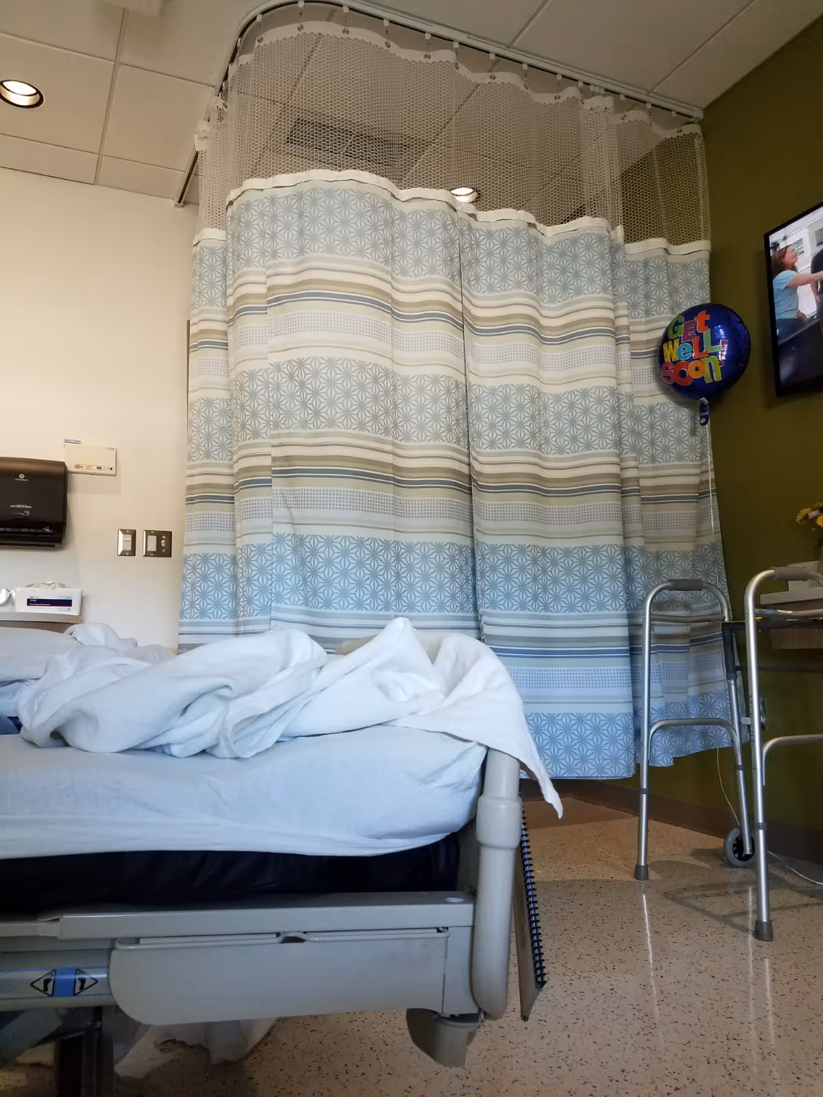 Hospital bed in a patient room with blue patterned privacy curtain and walker nearby.