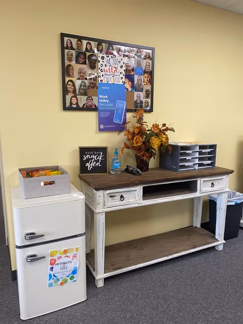 Rustic white console with a mini-fridge, snacks, flowers, and a photo collage on a yellow wall.