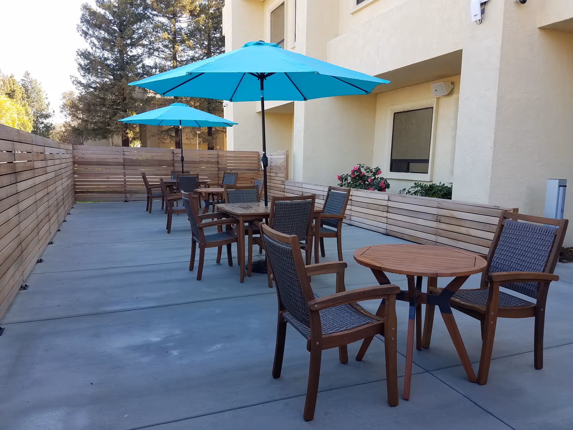 An outdoor patio with wooden tables, chairs, and teal umbrellas beside a beige building.