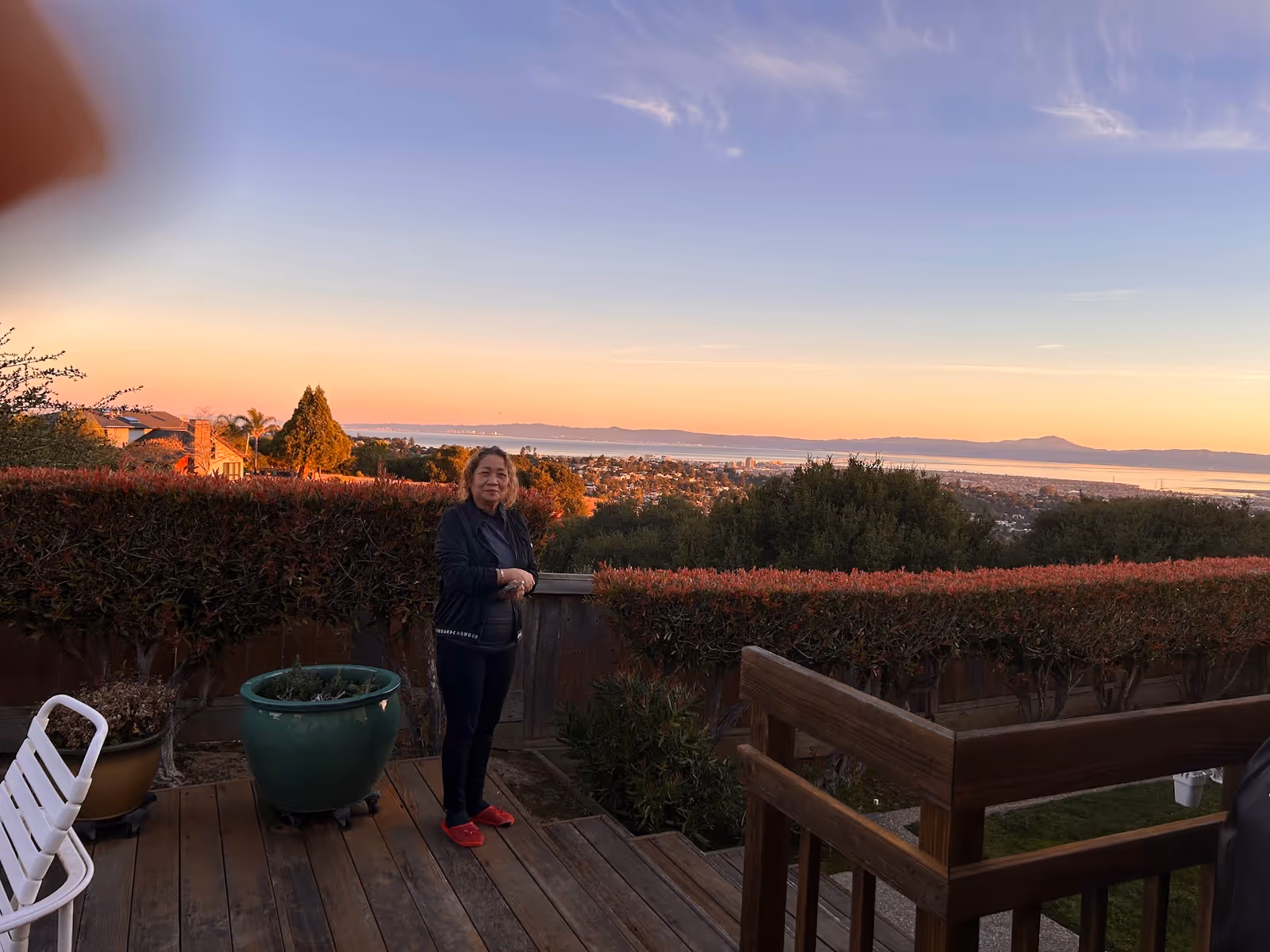 A person stands on a wooden deck with planters, overlooking a sunset coastal landscape.