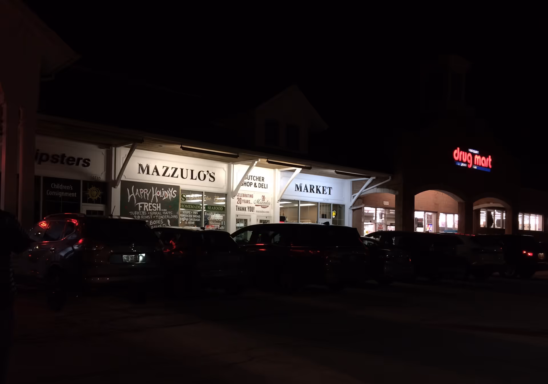 Nighttime storefronts with a lit Discount Drug Mart sign and parked cars in front.