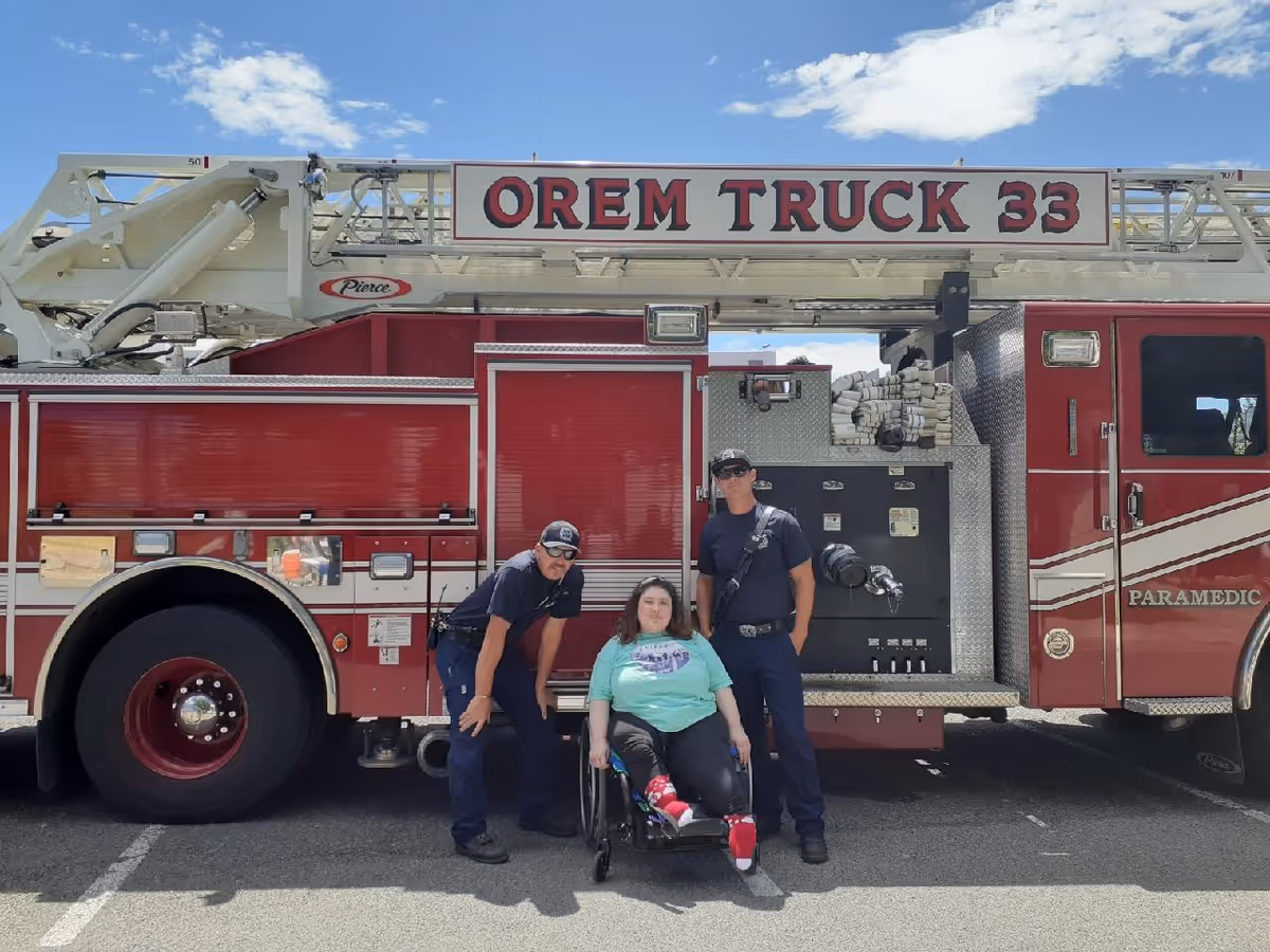 Three people, including two firefighters and a woman in a wheelchair, pose beside a fire truck.