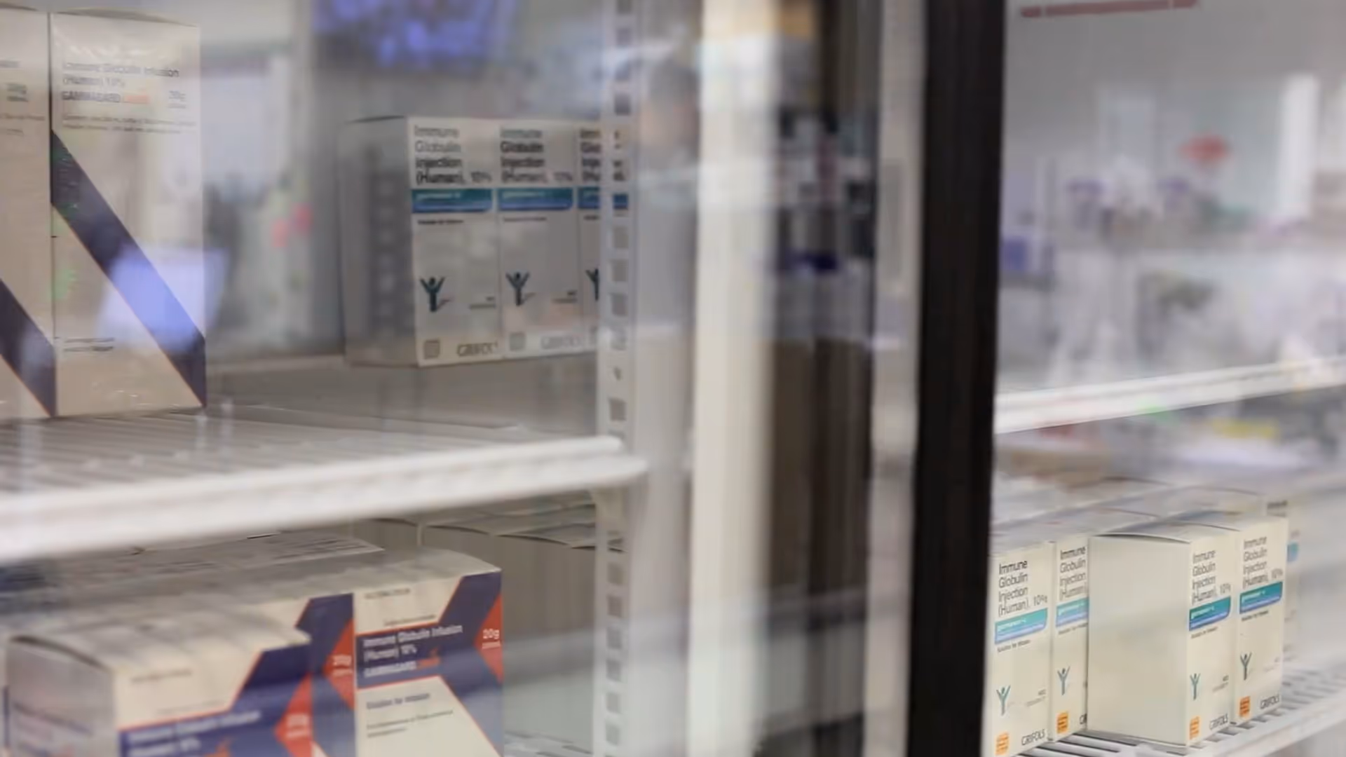Glass-front refrigerator stocked with boxed medications on metal shelves in a medical storage area.