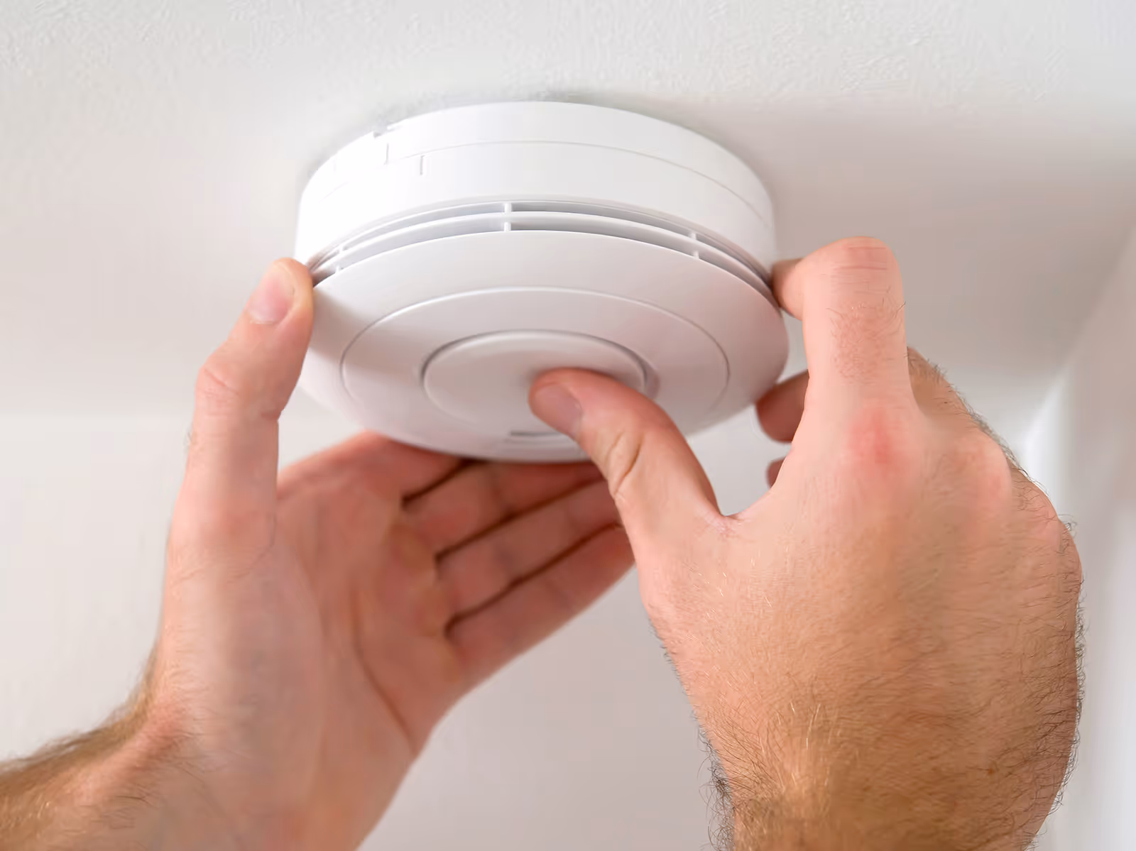 A pair of hands installs a round smoke detector on a white ceiling.