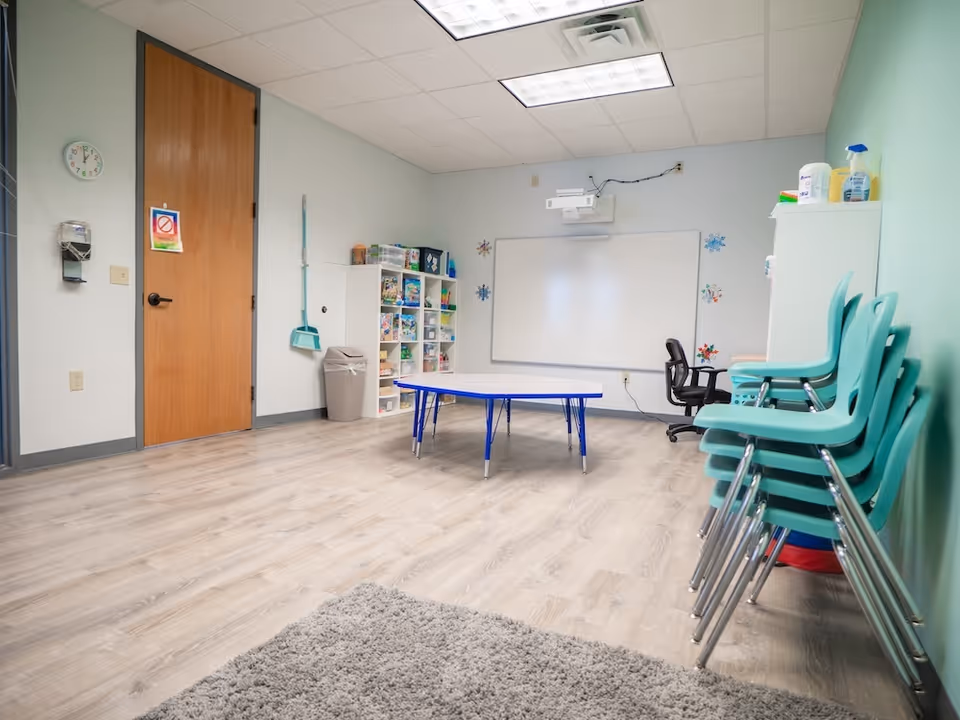 A bright classroom-style activity room with a whiteboard, shelves, and teal stacked chairs.